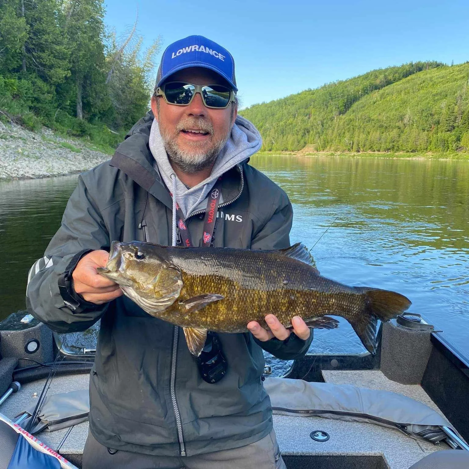A man holding a large fish on a boat in a river with forested hills in the background.