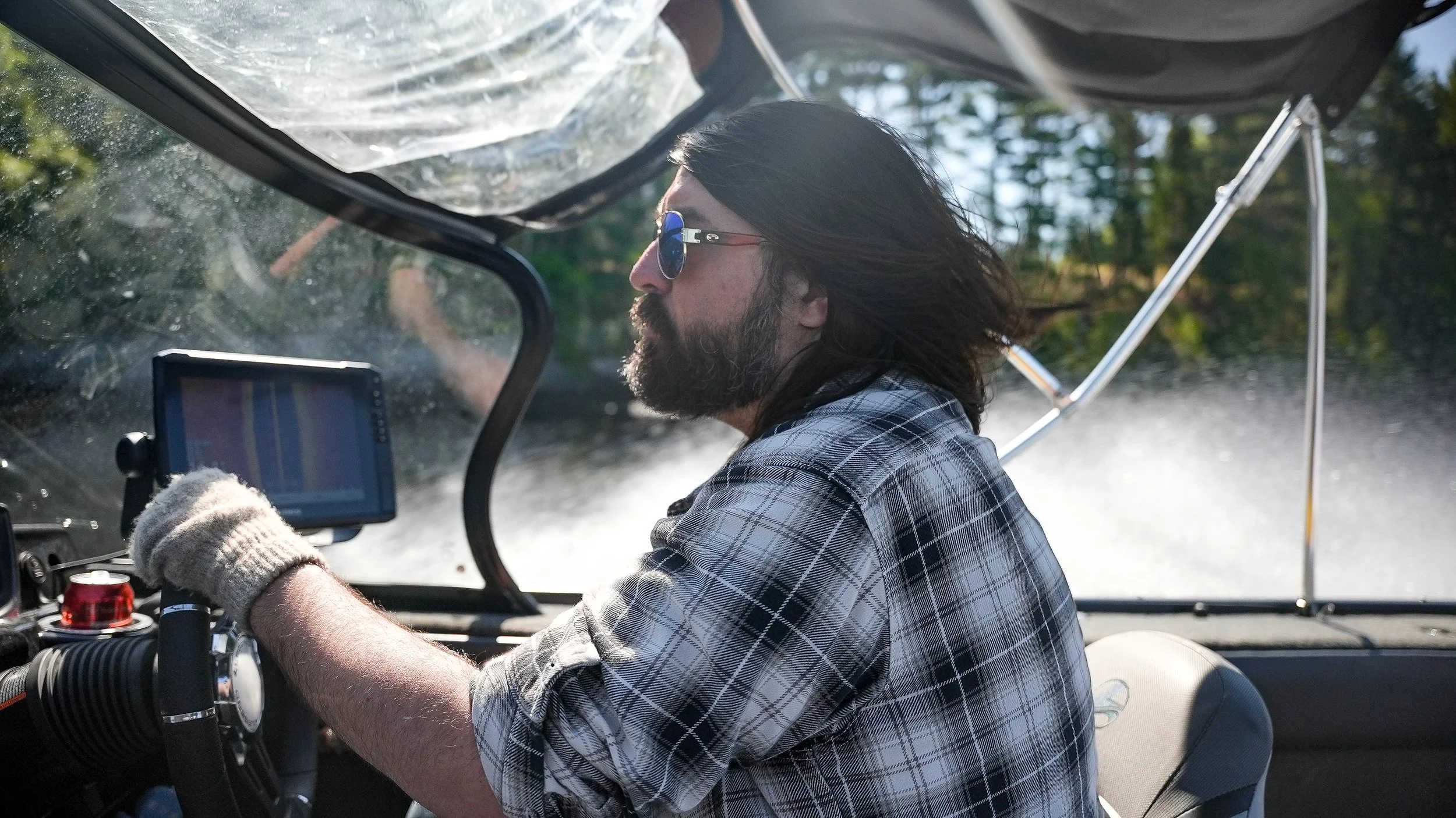 A man with long dark hair, a beard, wearing sunglasses, a plaid shirt, and a glove on his right hand, driving a boat on a sunny day with trees in the background.