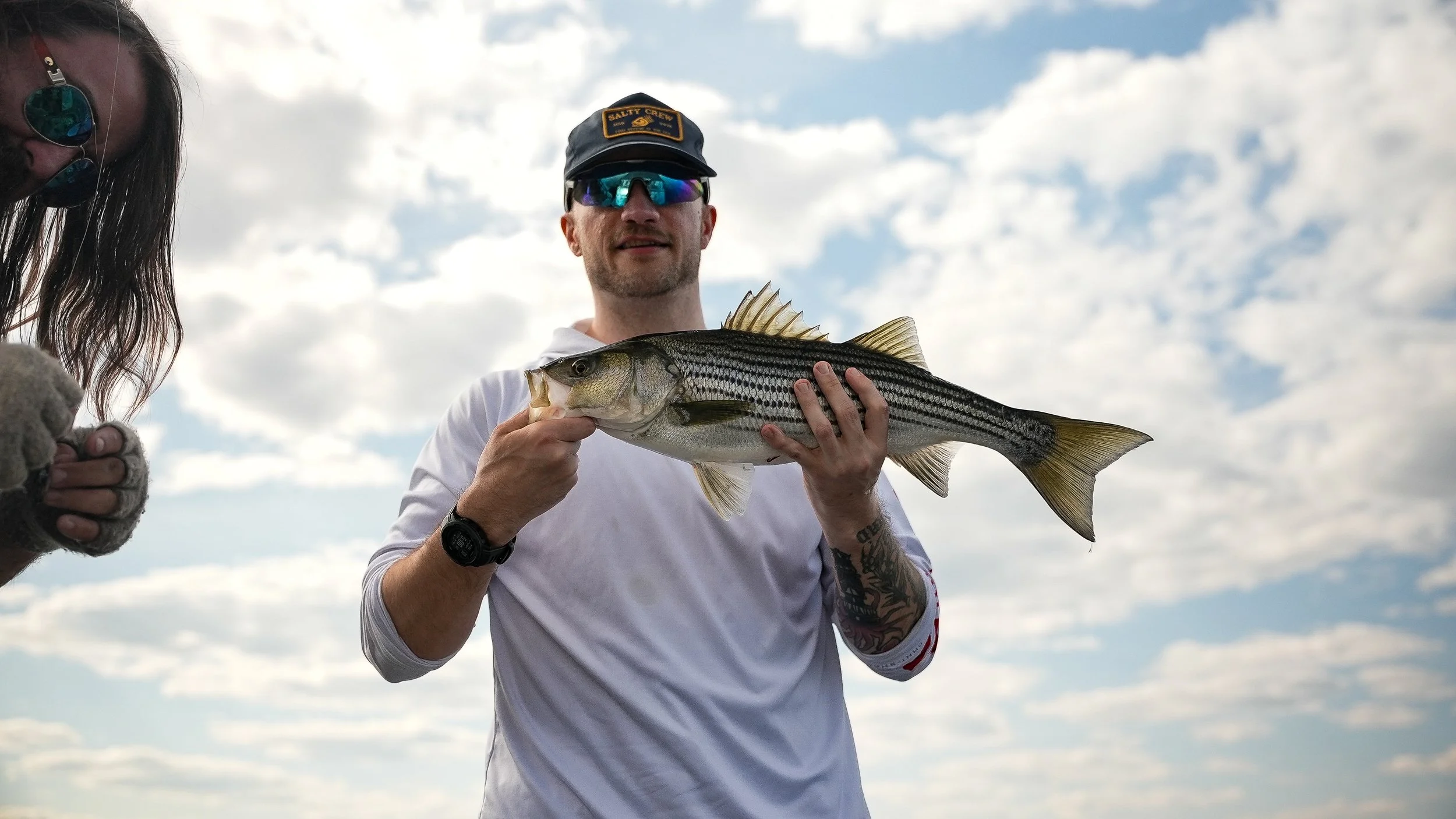 Man holding a freshly caught striped bass fish outdoors against a cloudy sky, wearing sunglasses, a cap, and a watch.