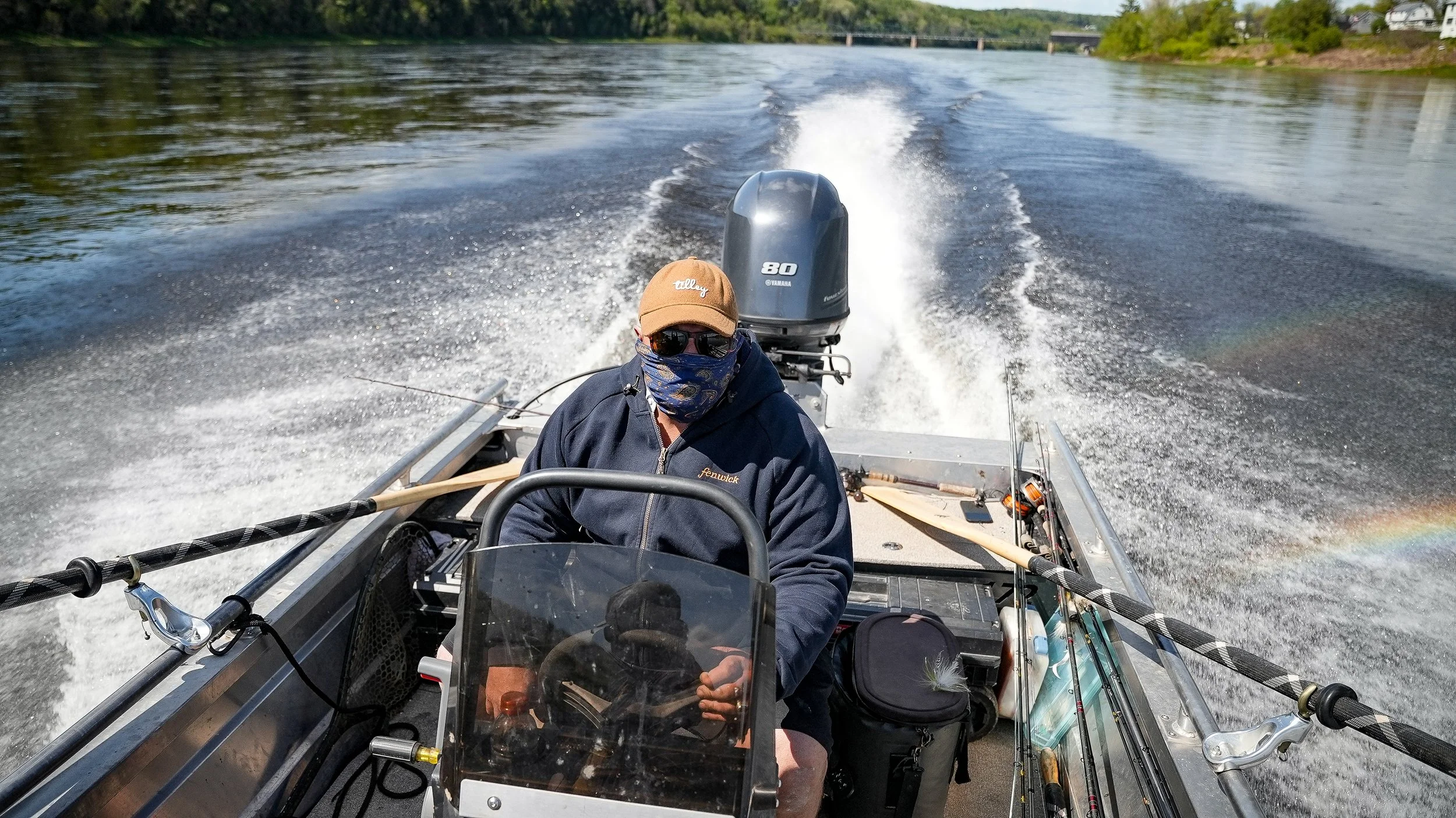 A person wearing sunglasses, a brown cap, and a face mask is steering a speedboat on a river, with a Yamaha outboard motor at the back. The boat is moving swiftly, creating waves and a rainbow on the water's surface.