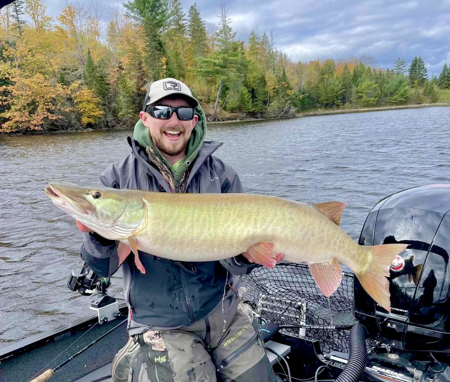 Man in outdoor clothing on boat holding large fish, with trees and lake in background.