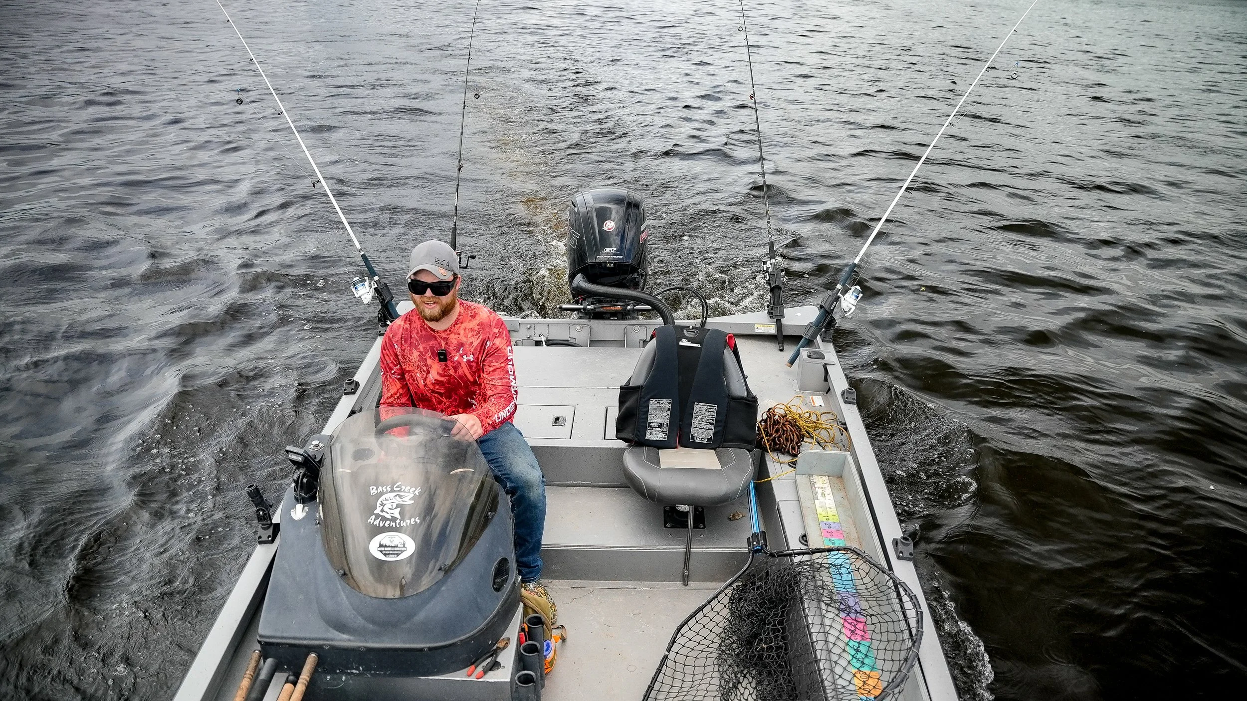 A man with sunglasses and a red shirt sitting at the helm of a boat on a body of water, with fishing rods on each side, a motor at the back, and various fishing equipment on the boat.