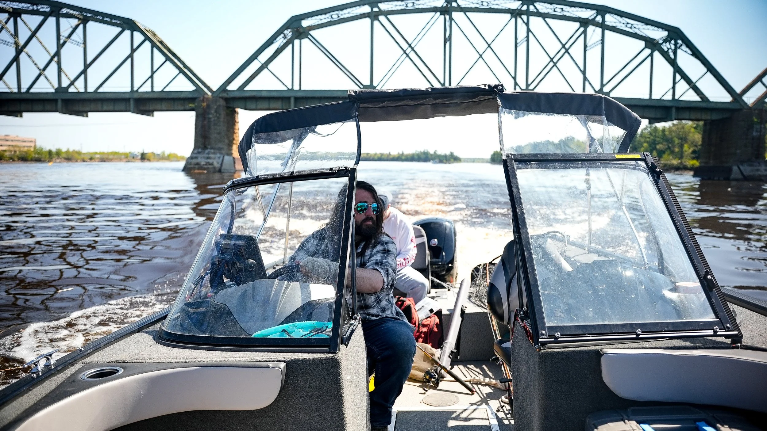 A man with sunglasses and a checkered shirt driving a small motorboat on a body of water, with a bridge in the background and another person behind him. The boat has a clear windshield and appears to be moving quickly.
