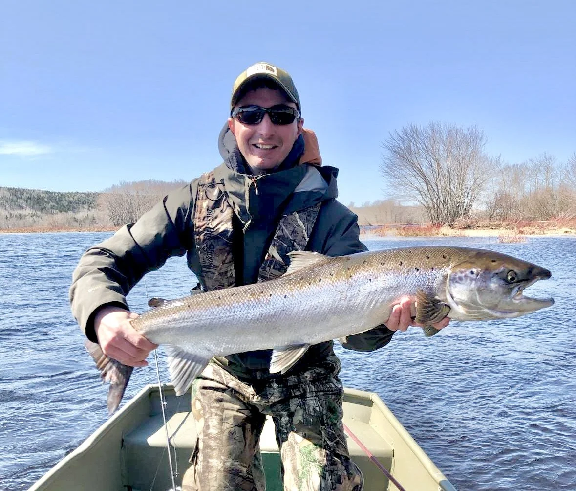 Man in camouflage jacket and baseball cap holding a large fish on a boat in a lake during daytime.