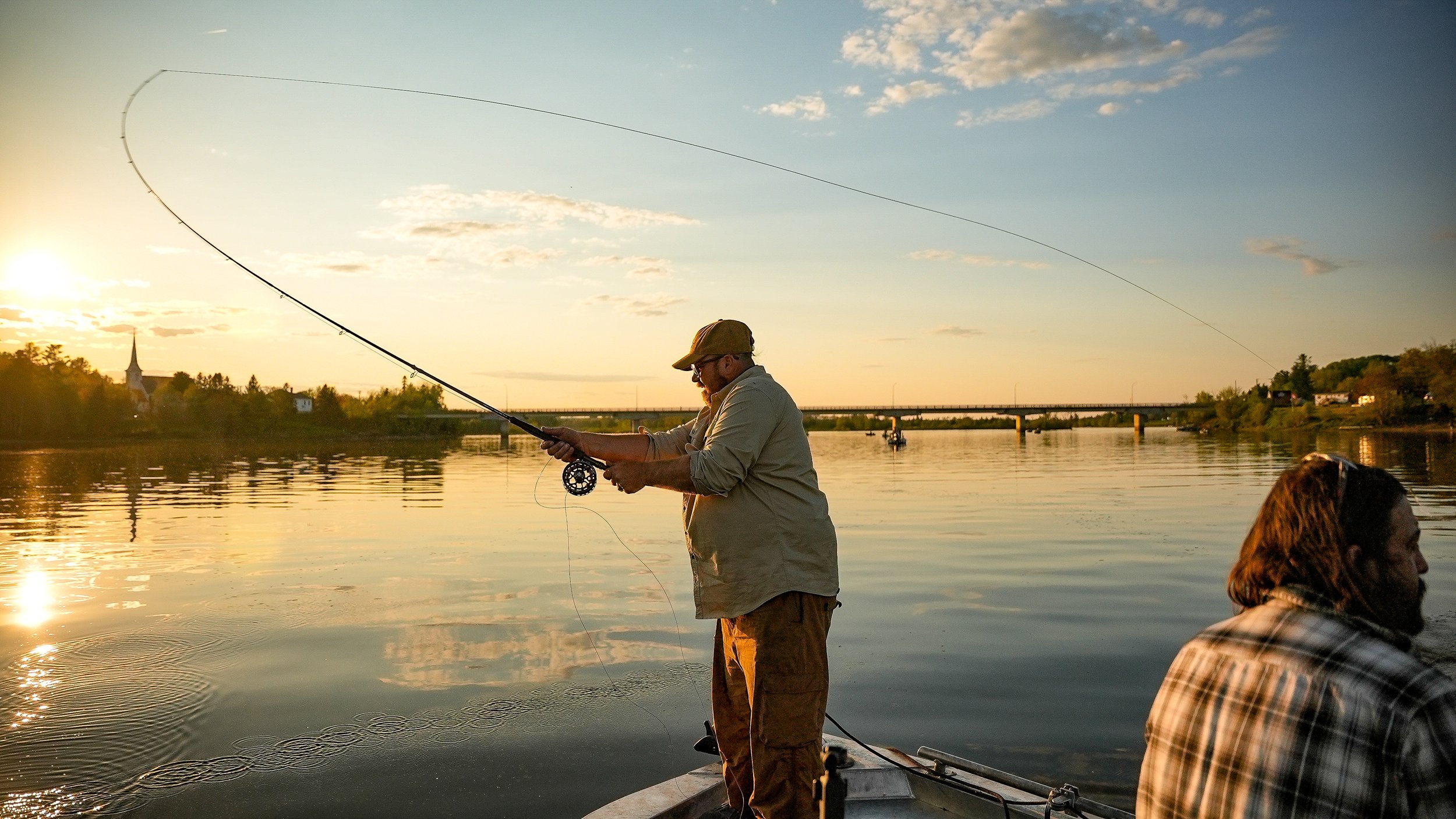 Two men fishing from a boat on a river during sunset, with trees and a bridge in the background.