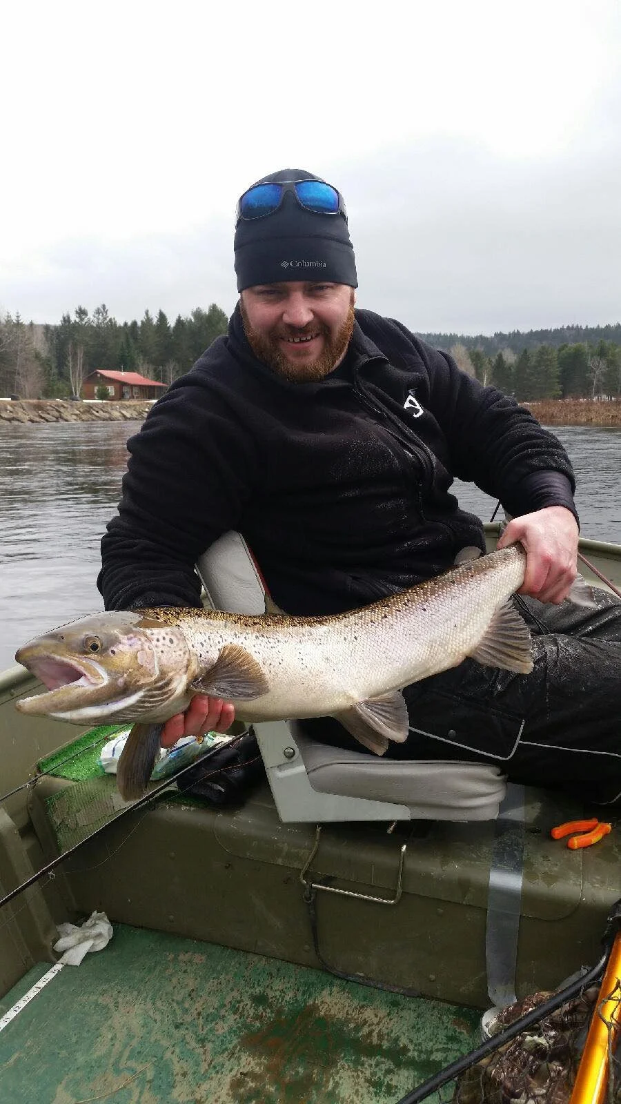 A man sitting in a boat on a lake, holding a large fish with a wide open mouth, wearing a black jacket and winter hat with sunglasses on top, with trees and a house visible in the background.