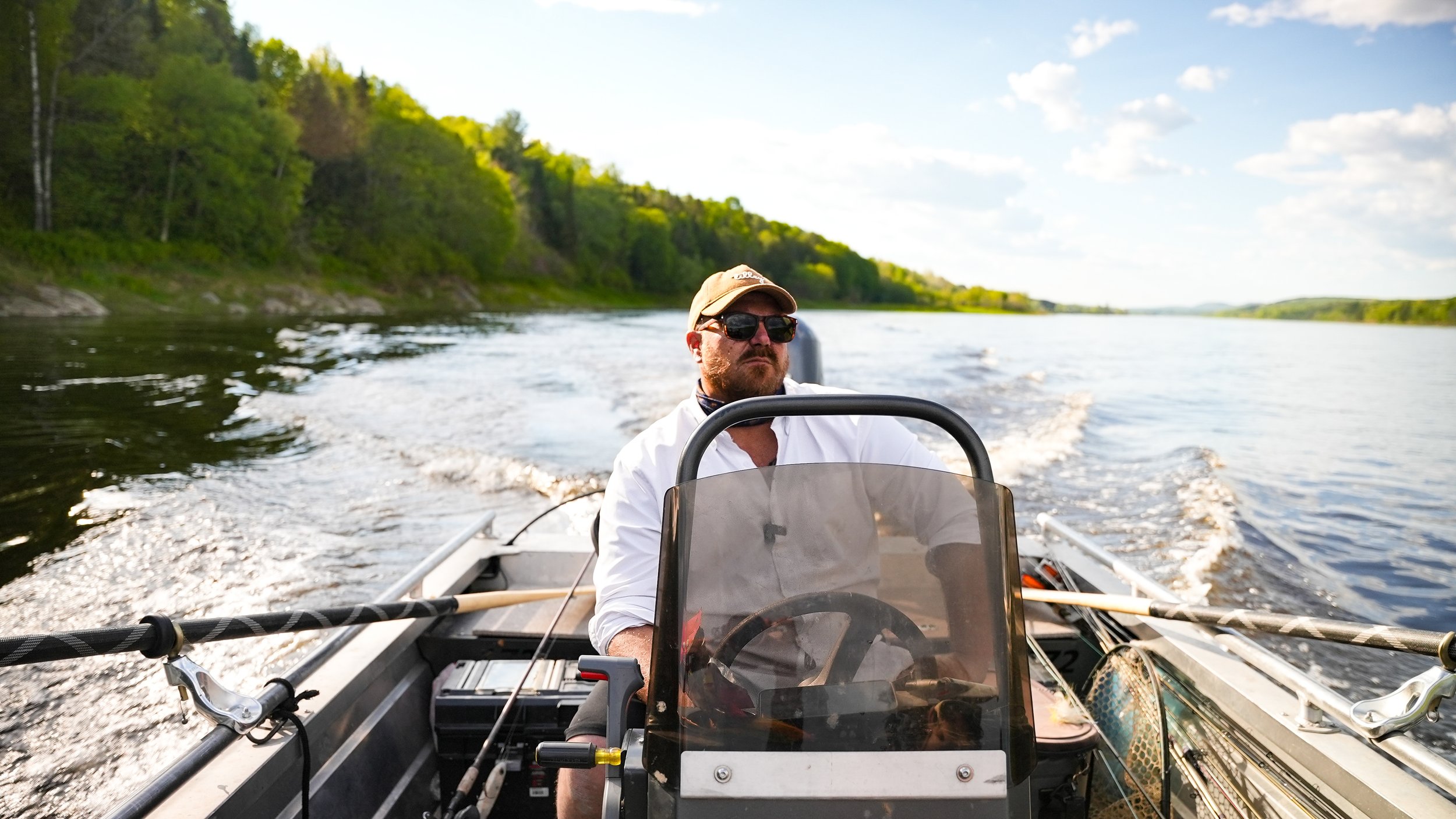 A man wearing sunglasses, a hat, and a white shirt driving a boat on a river during daytime, with lush green trees on the riverbank and partly cloudy sky.