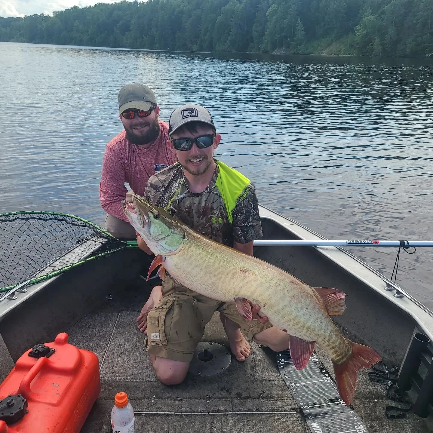 Two men on a boat with a large fish, possibly a walleye, in a lake with trees in the background