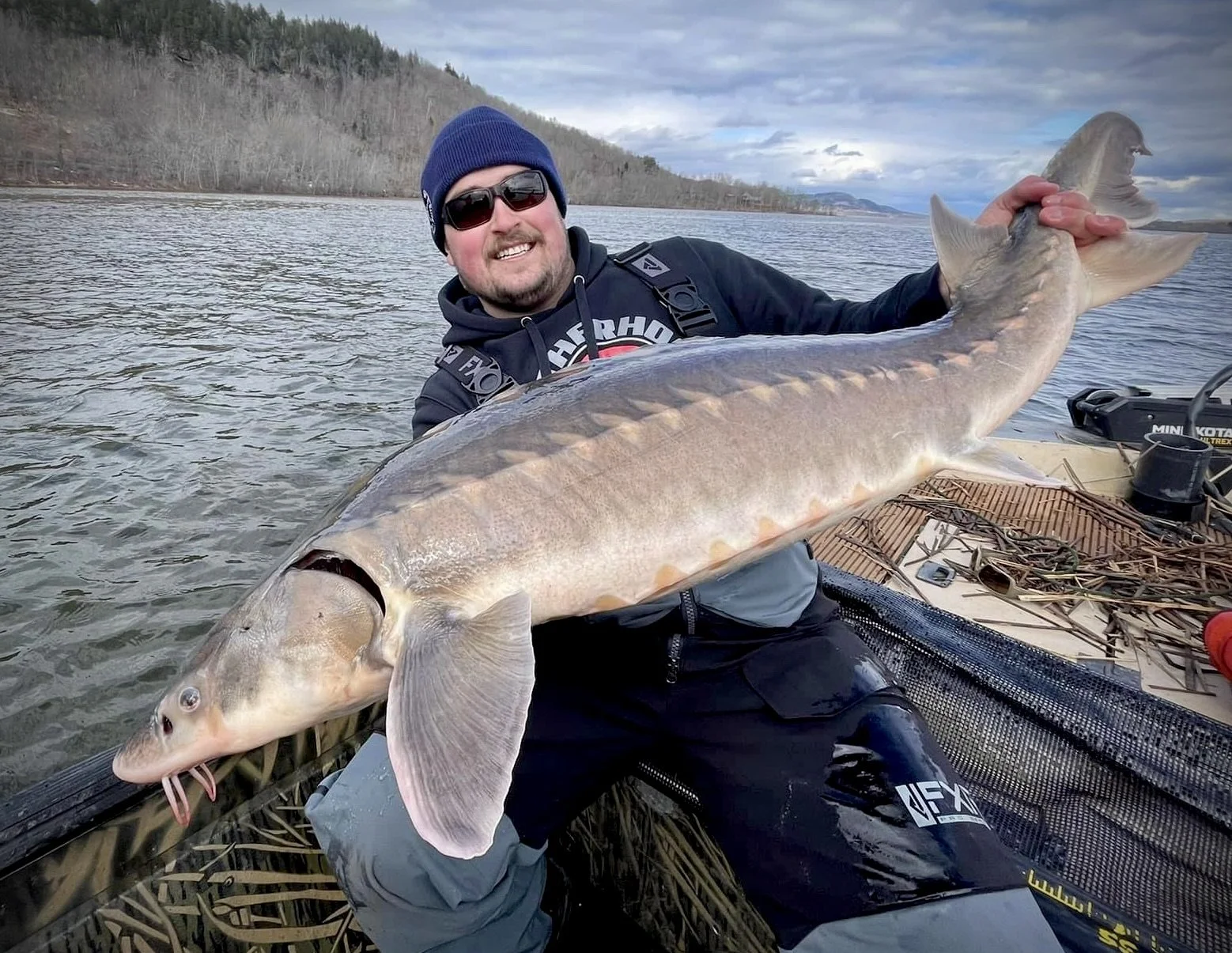 A man dressed in orange fishing gear sits in a boat, holding a large fish with a striped pattern. The background shows a calm lake, a hillside, and a partly cloudy sky.