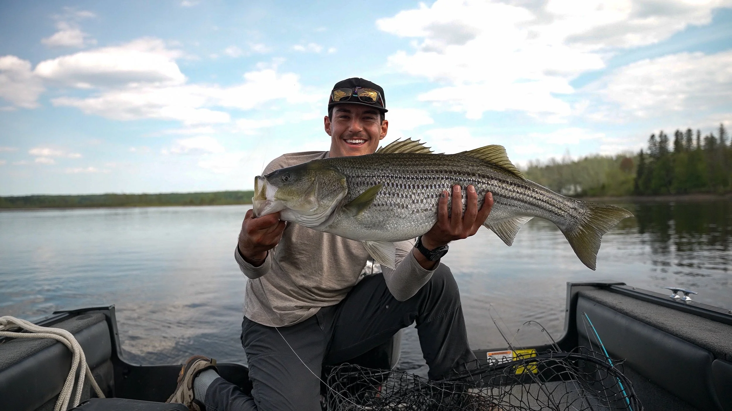 A man smiling and holding a large fish on a boat in a lake with trees in the background and cloudy sky.
