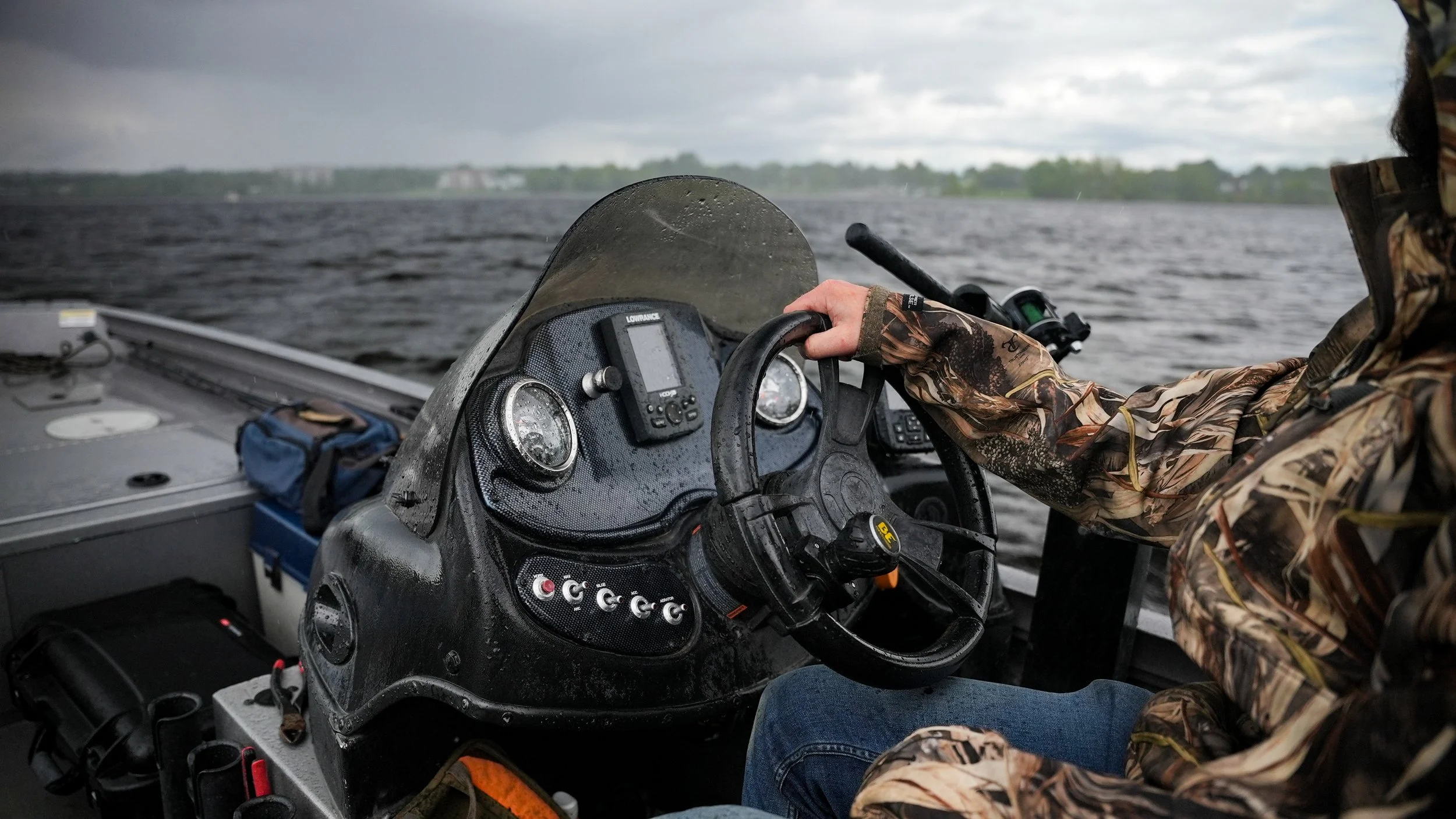 Person wearing camouflage jacket steering a boat on a large body of water, cloudy sky in the background.