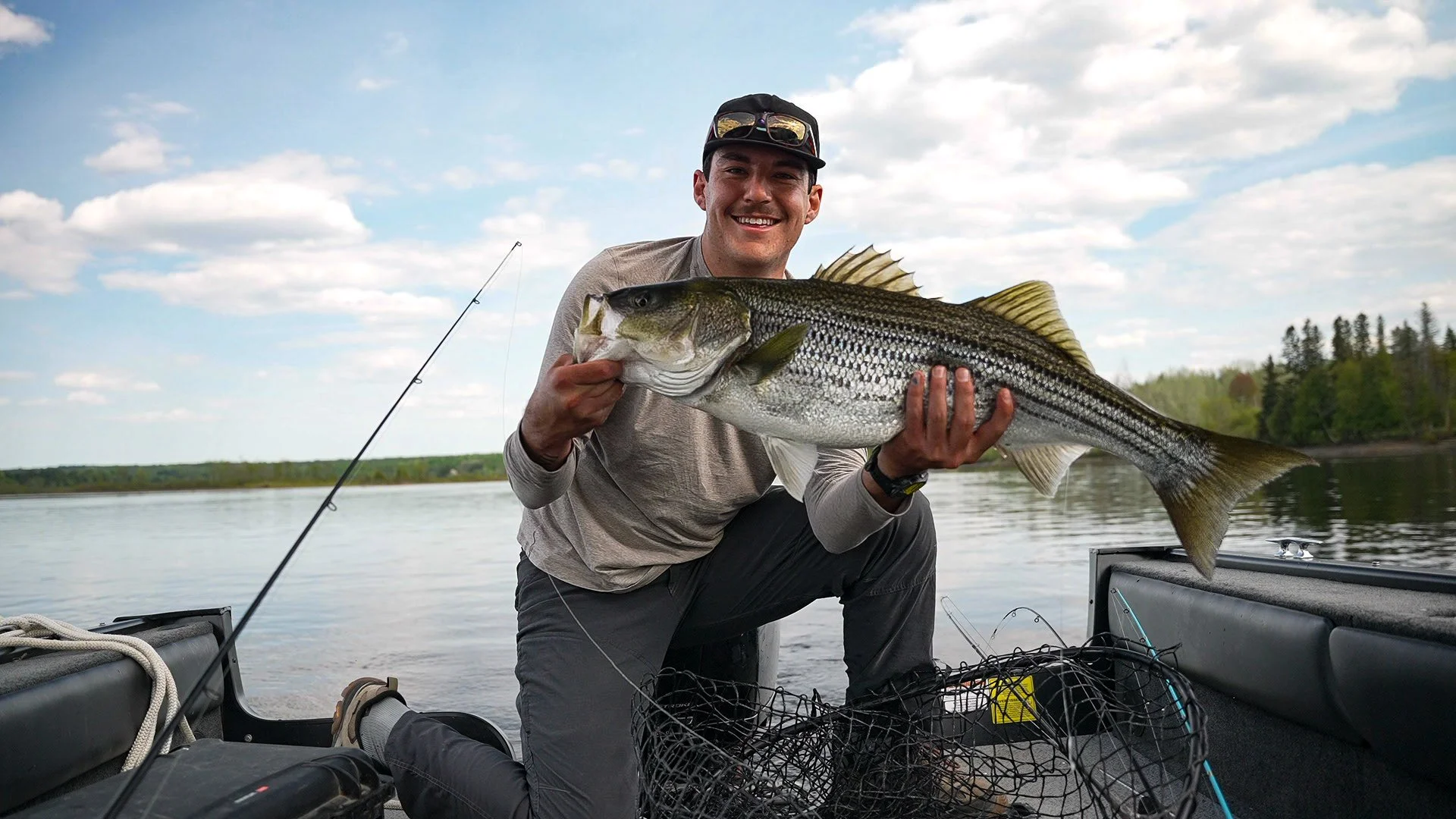 Man holding a large striped bass fish on a boat on a calm lake with trees in the background.