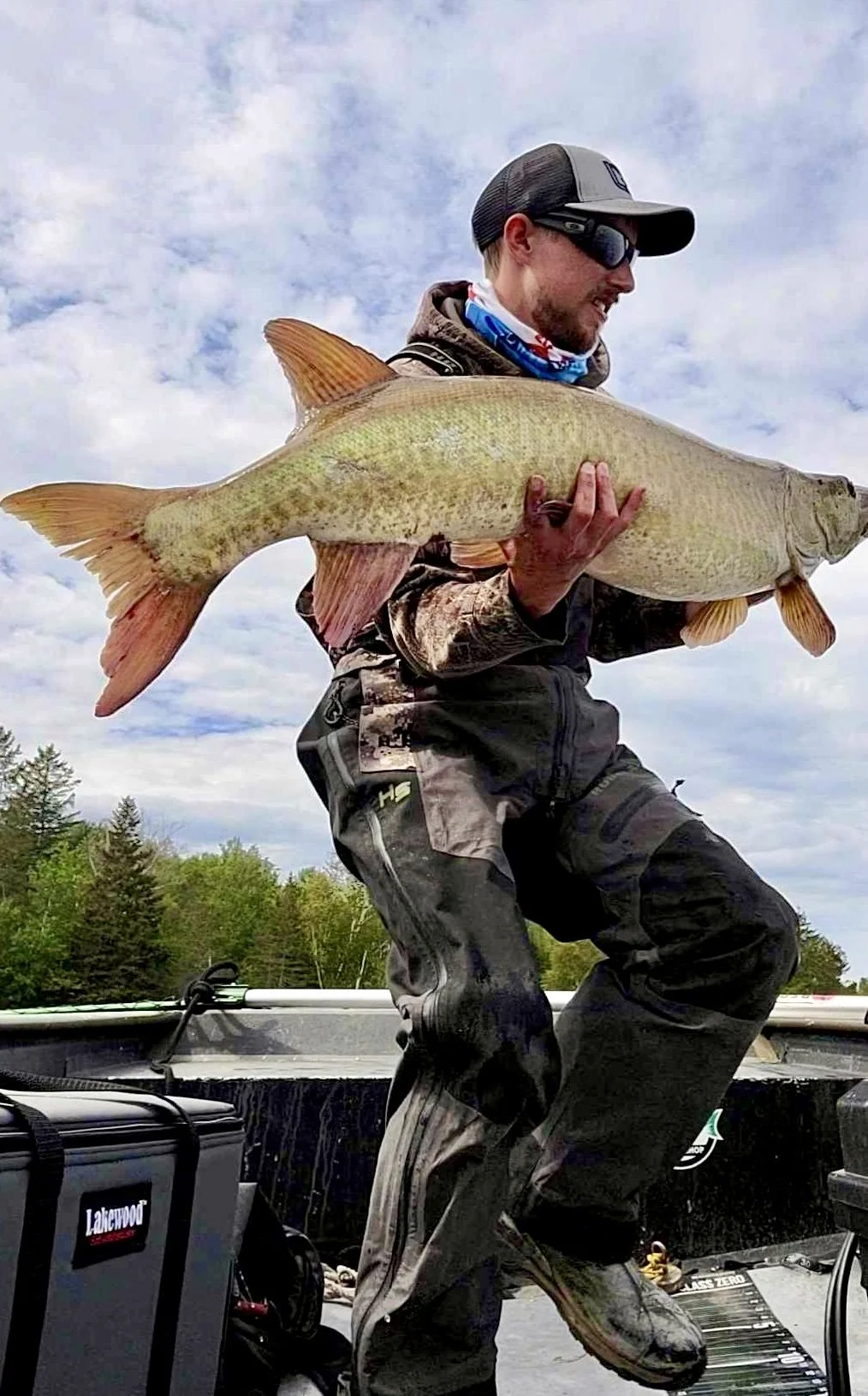 Man in outdoor gear holding a large fish, standing on a boat with trees and cloudy sky in the background.