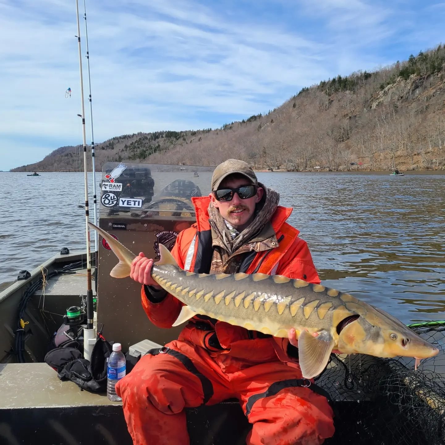 A man dressed in orange fishing gear sits in a boat, holding a large fish with a striped pattern. The background shows a calm lake, a hillside, and a partly cloudy sky.