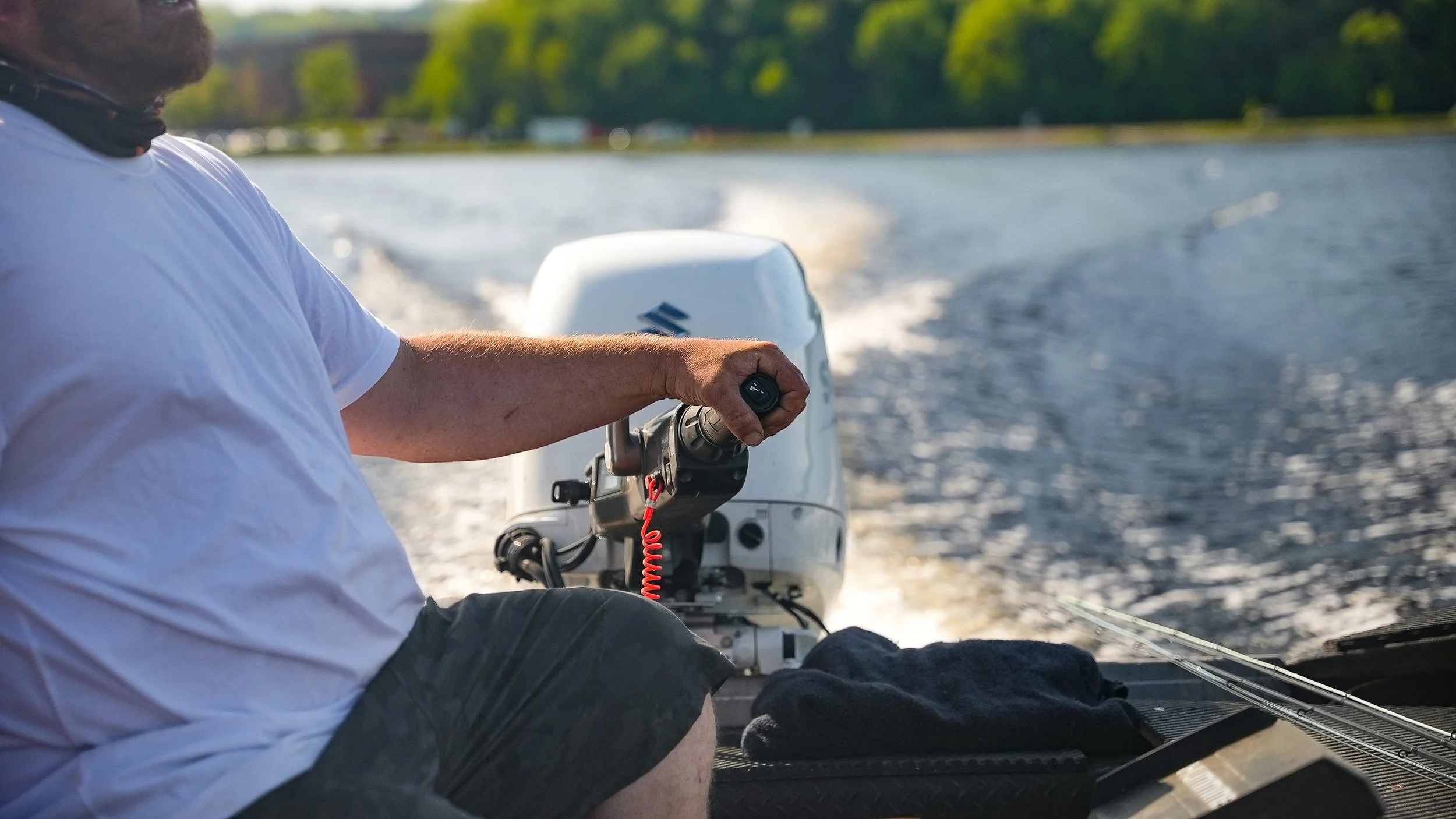 Person steering a boat with a white outboard motor on a lake, wearing a white shirt and shorts, with a towel and equipment in the boat.