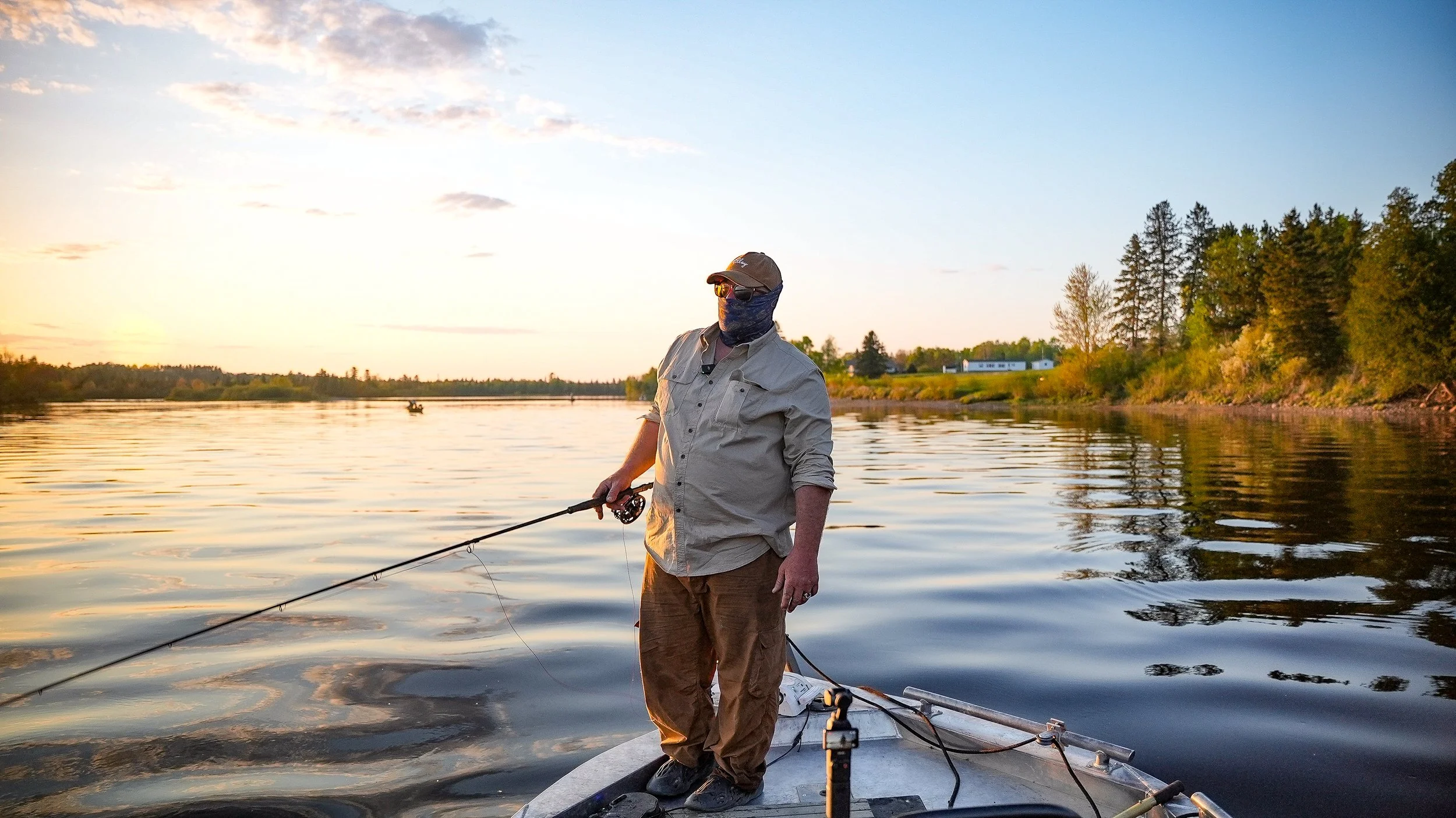 Person fishing from a boat on a calm river during sunset, with lush trees and a blue sky in the background.