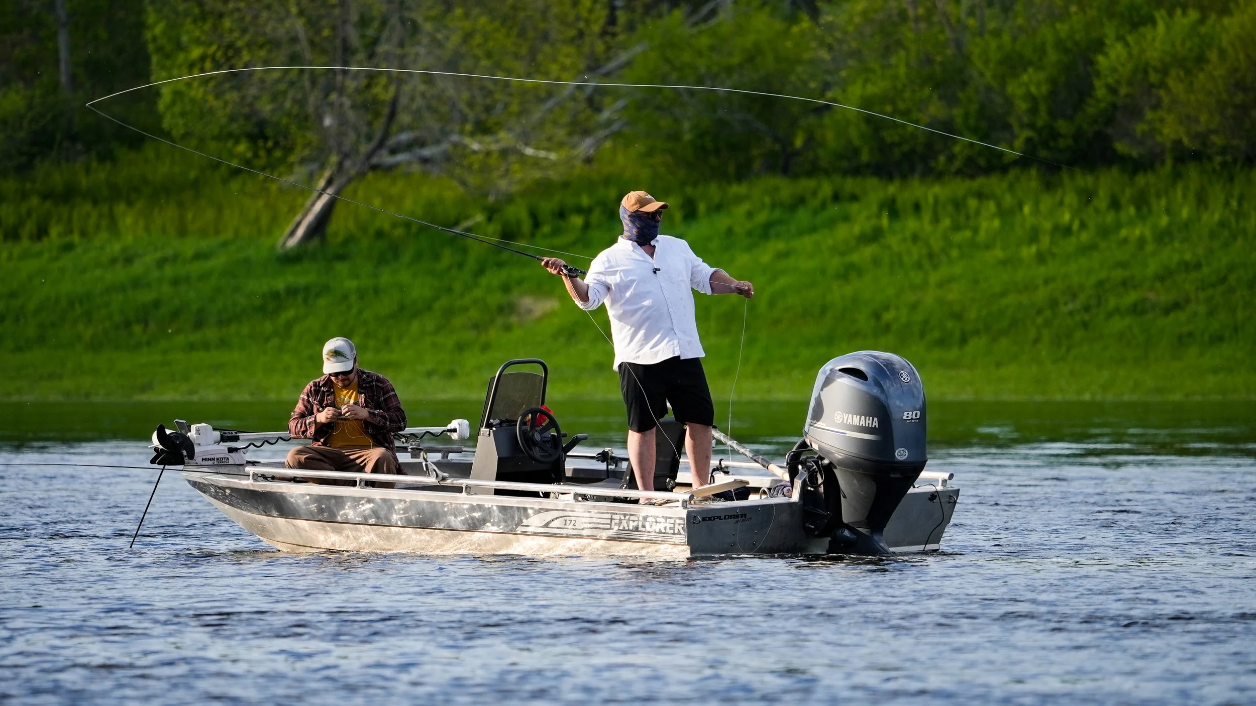 Two men fishing on a boat in a lake with green trees in the background. One man is standing and casting a fishing line, wearing a white shirt, black shorts, a hat, and a face covering. The other man is sitting, looking at his phone, wearing a plaid s