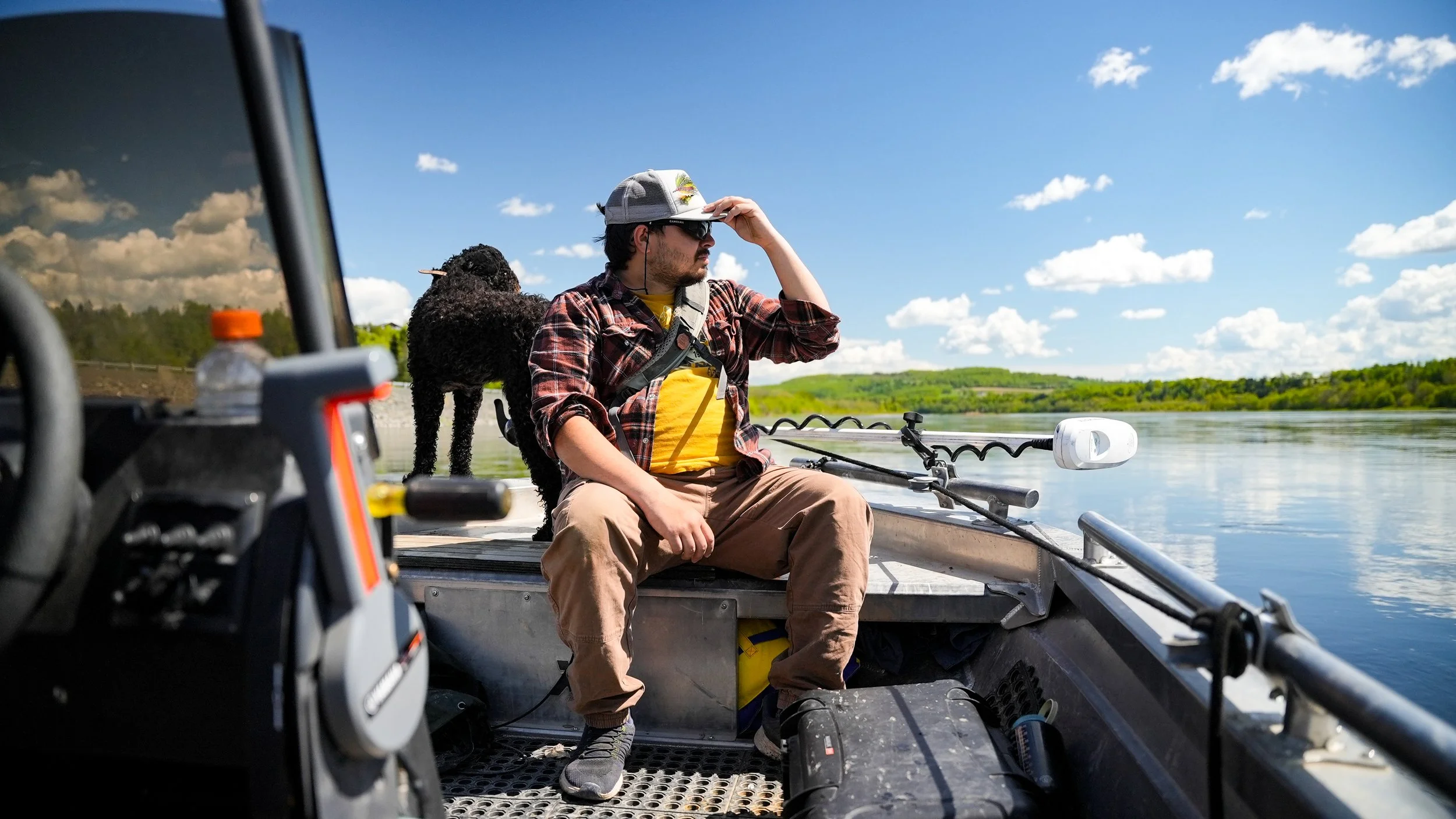 Man sitting on a boat by a lake with a dog, wearing a hat and sunglasses, holding his hand to his forehead, with green trees and blue sky in the background.