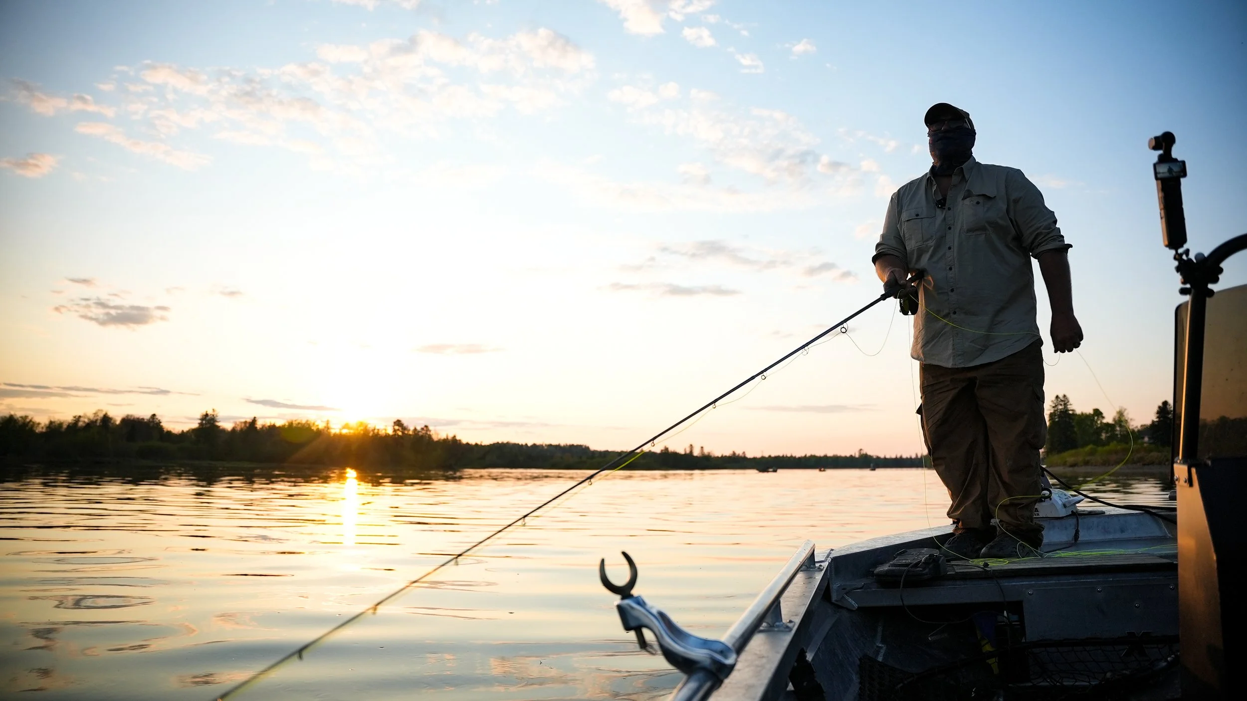 A person fishing on a boat during sunset on a calm lake, with a silhouette of trees in the background.
