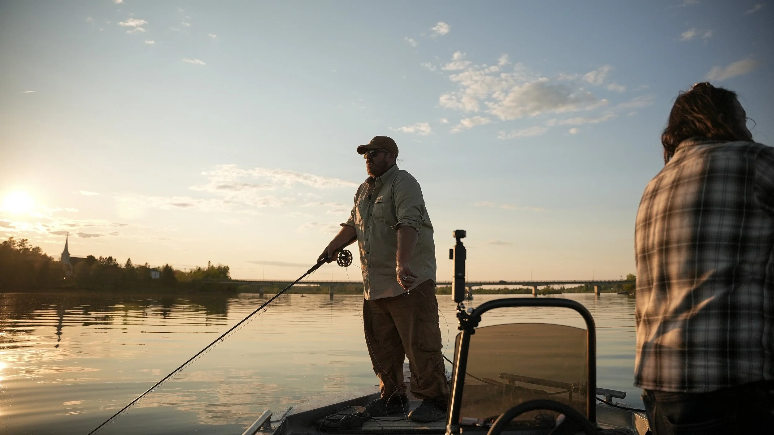 A man and woman fishing from a boat on a river during sunset, with a church steeple in the background.