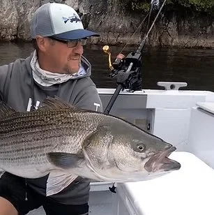 Man in a gray jacket, sunglasses, and a white and gray cap fishing on a boat, holding a large fish.