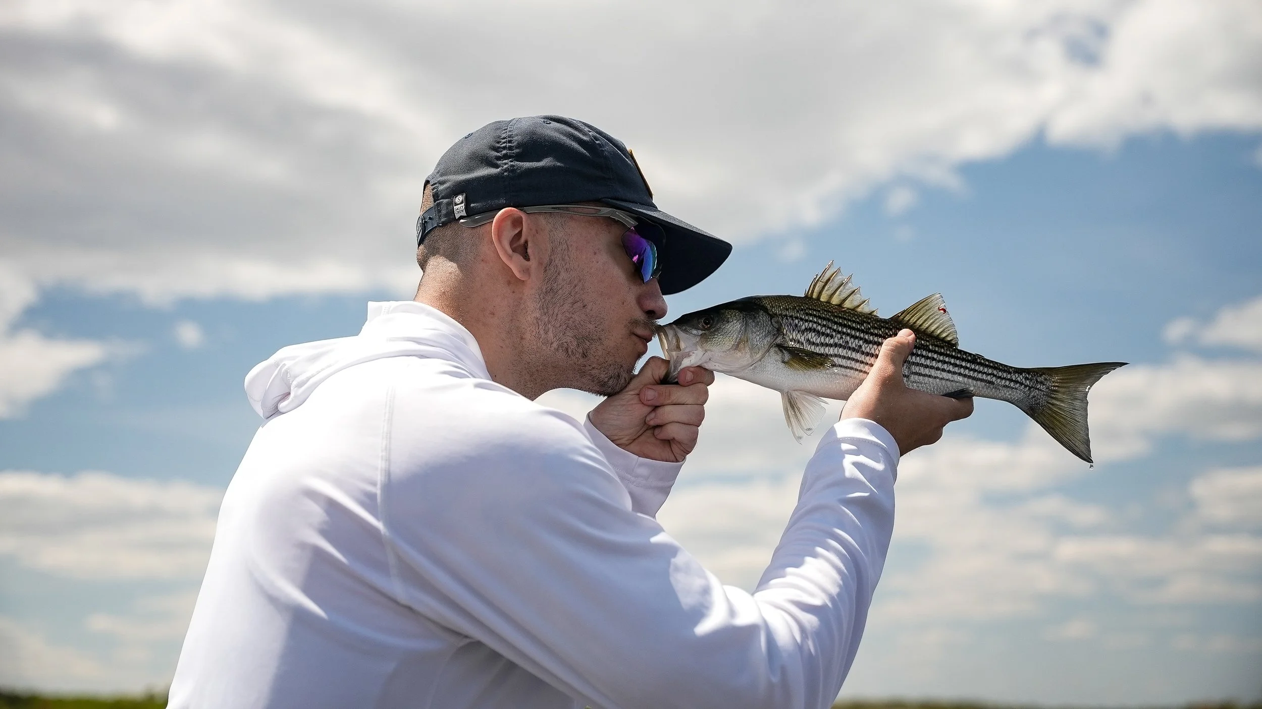 A man wearing sunglasses and a black cap kisses a fish he is holding, outdoors with a cloudy sky.