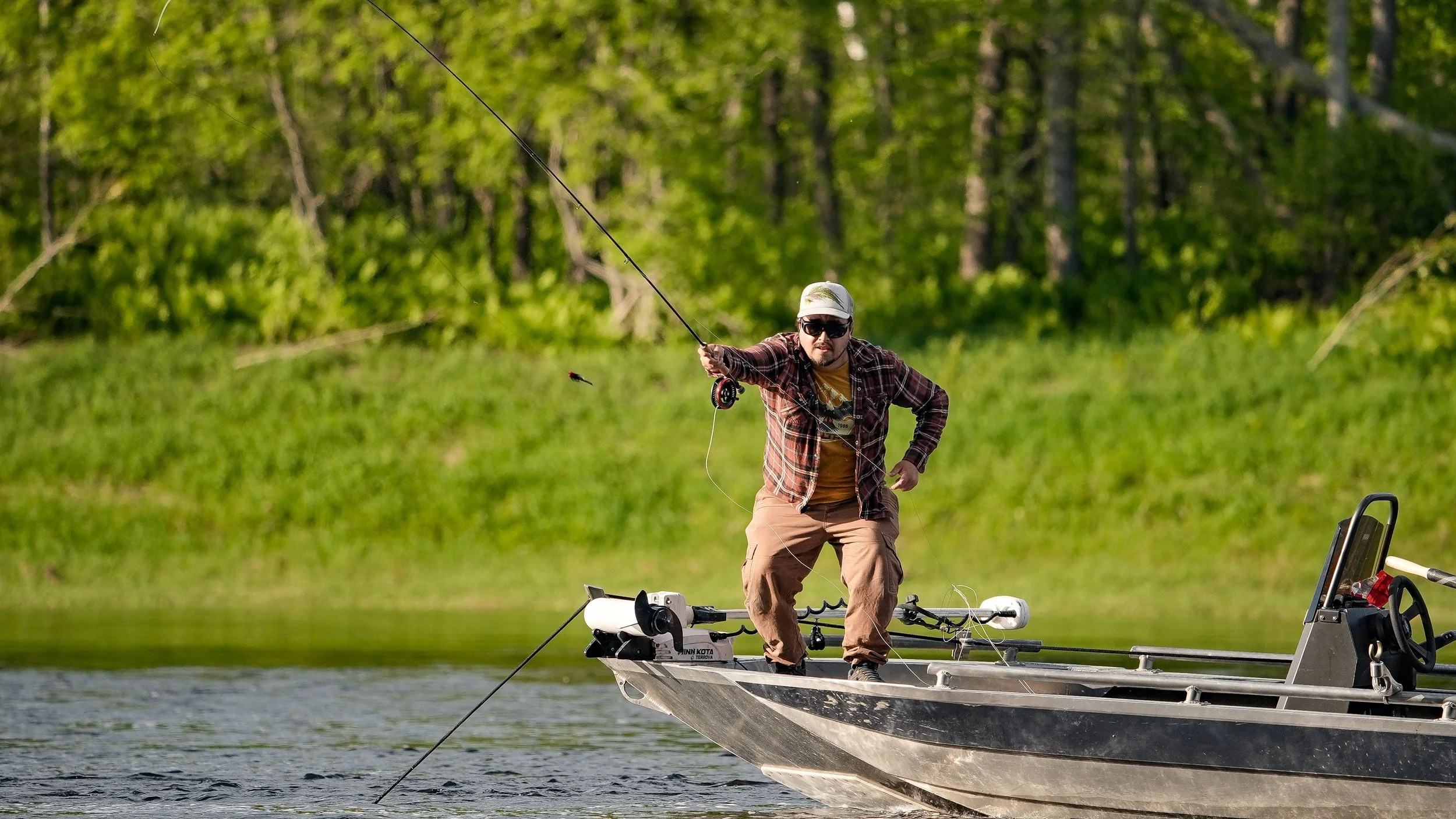 Man in sunglasses, cap, and plaid shirt fishing from a boat on a lake with green trees in the background.