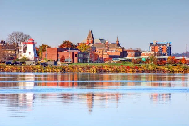 A cityscape along a river with historic buildings, trees with fall foliage, and a lighthouse in the distance under a clear blue sky.