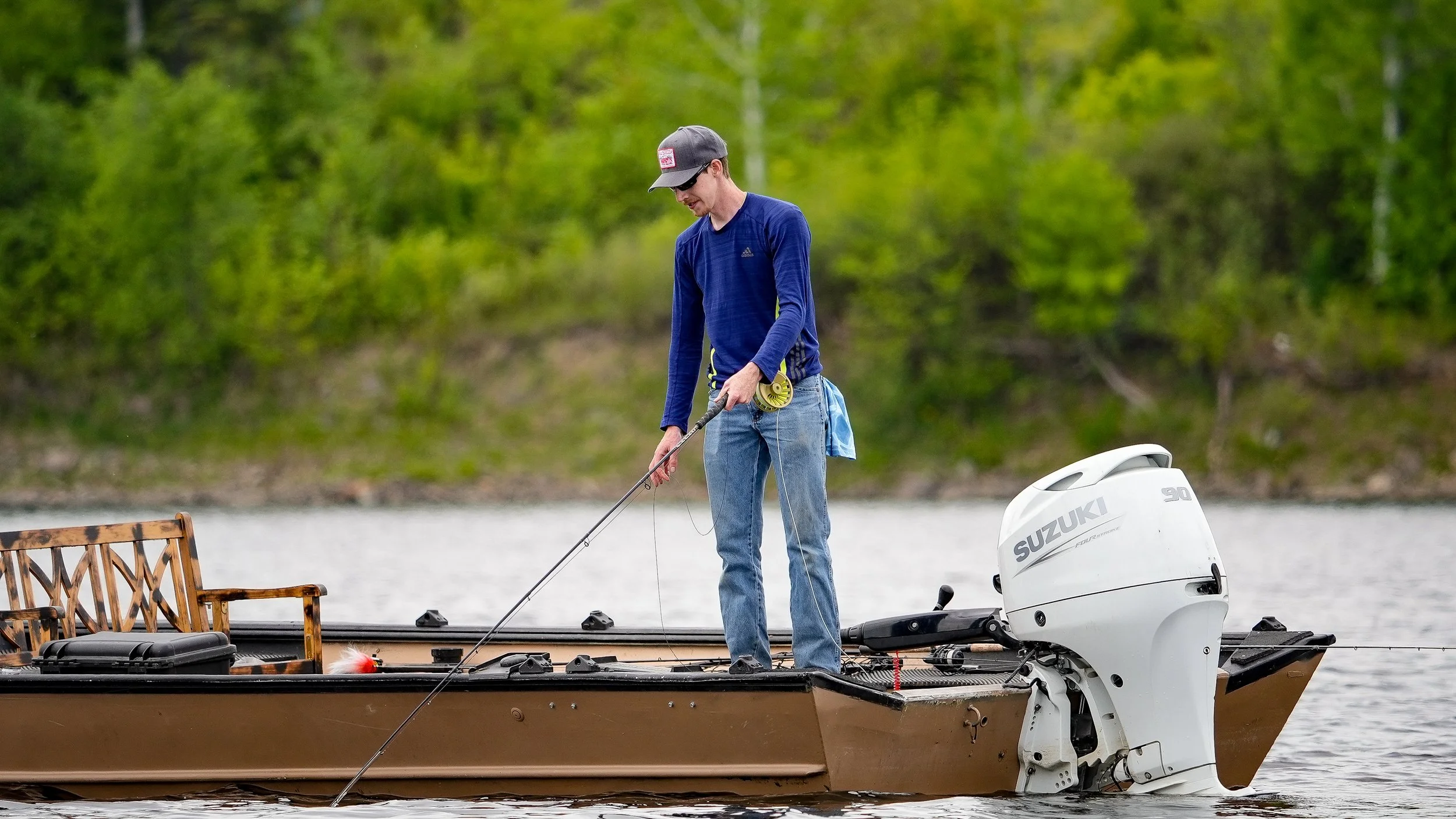 A man fishing on a boat with a Suzuki outboard motor, surrounded by a lake and green trees.