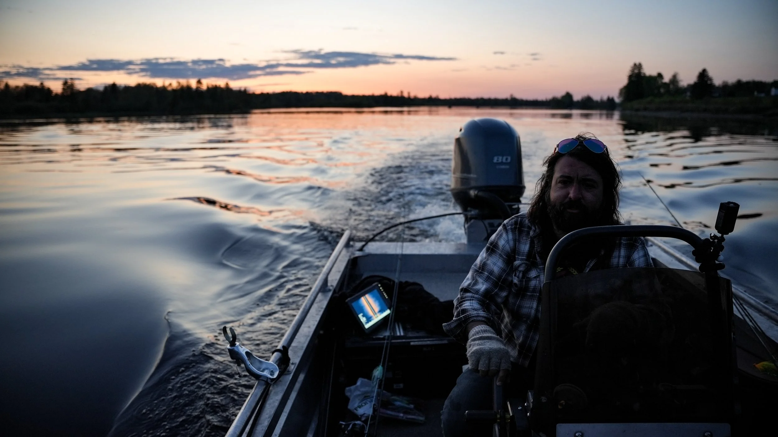 Man with long hair and beard wearing a plaid shirt and sunglasses on his head, sitting on a boat during sunset with calm water in the background, operating a motorboat.