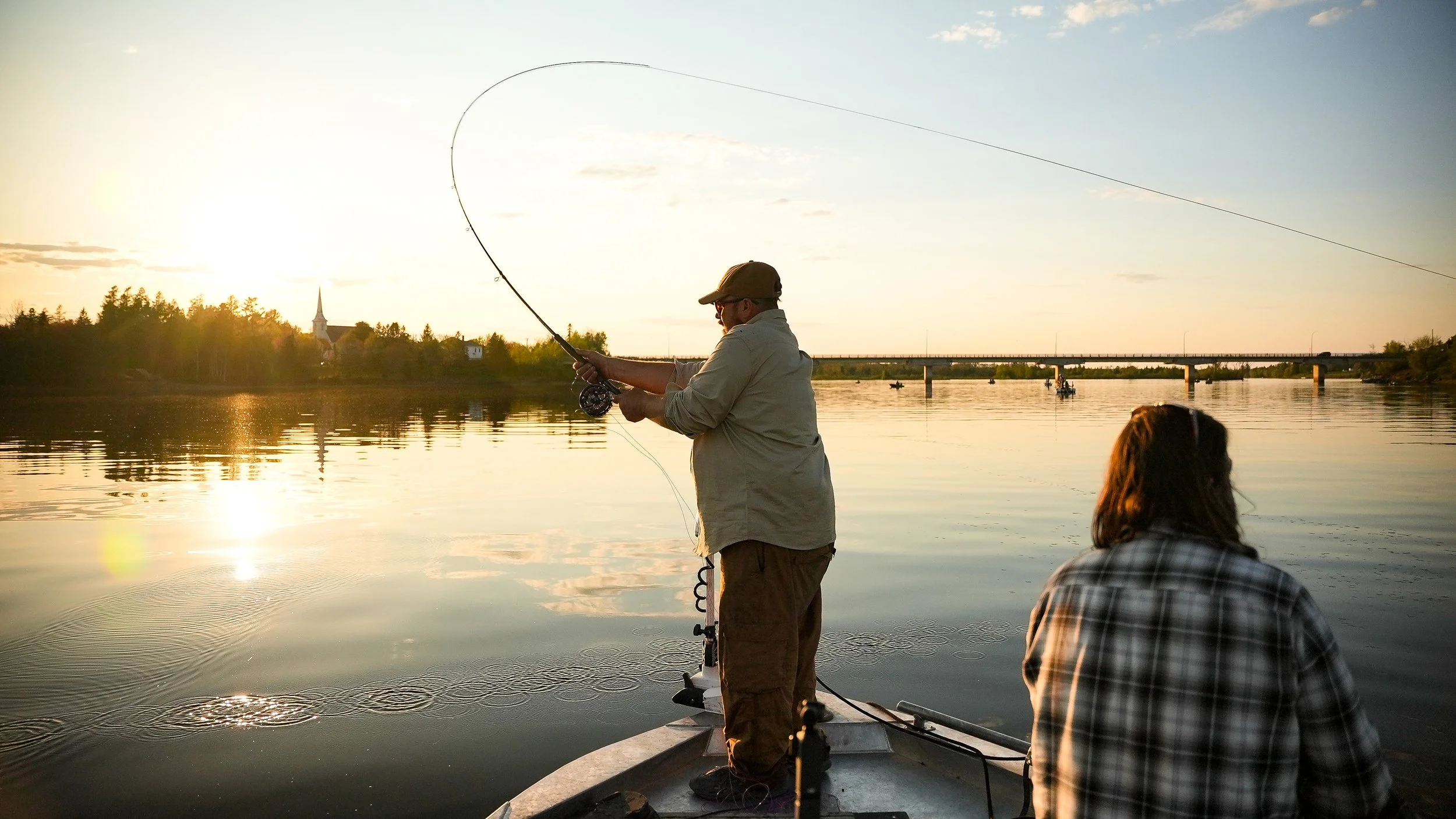 Two people fishing on a boat in a river during sunset, with one standing and casting a fishing line and the other sitting facing the water, and a bridge in the background.
