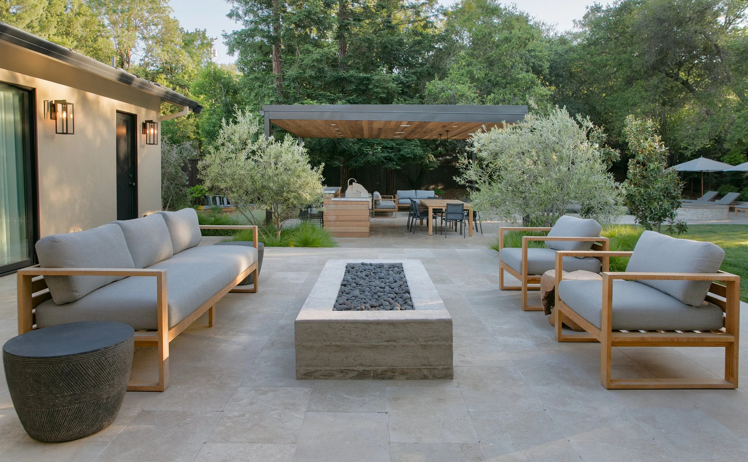 Modern outdoor patio with gray cushioned seating, a fire feature containing black stones, and a large shaded outdoor dining and lounging area with a built-in grill, surrounded by trees and greenery.