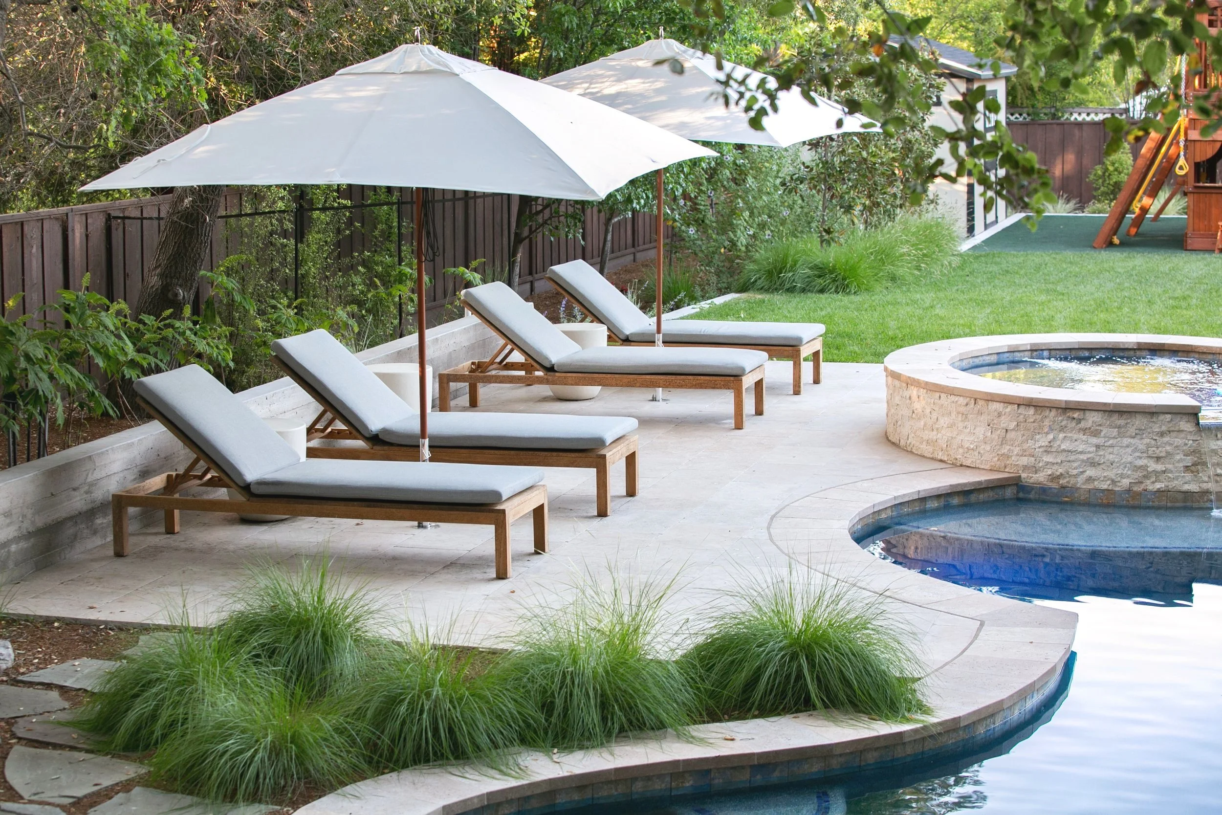 Outdoor backyard scene with three lounge chairs under white umbrellas, near a hot tub and swimming pool, surrounded by green grass, plants, and trees.