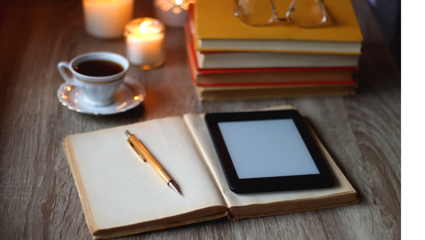 A wooden table with a white ceramic cup of black coffee on a matching saucer, lit candles, a closed notebook with a pen, an e-reader, and a stack of colorful books.