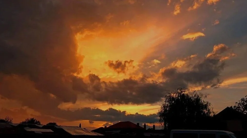 Dramatic orange sunset with dark clouds and golden light breaking through the sky above rooftops, symbolising emotional transformation and energetic balance.
