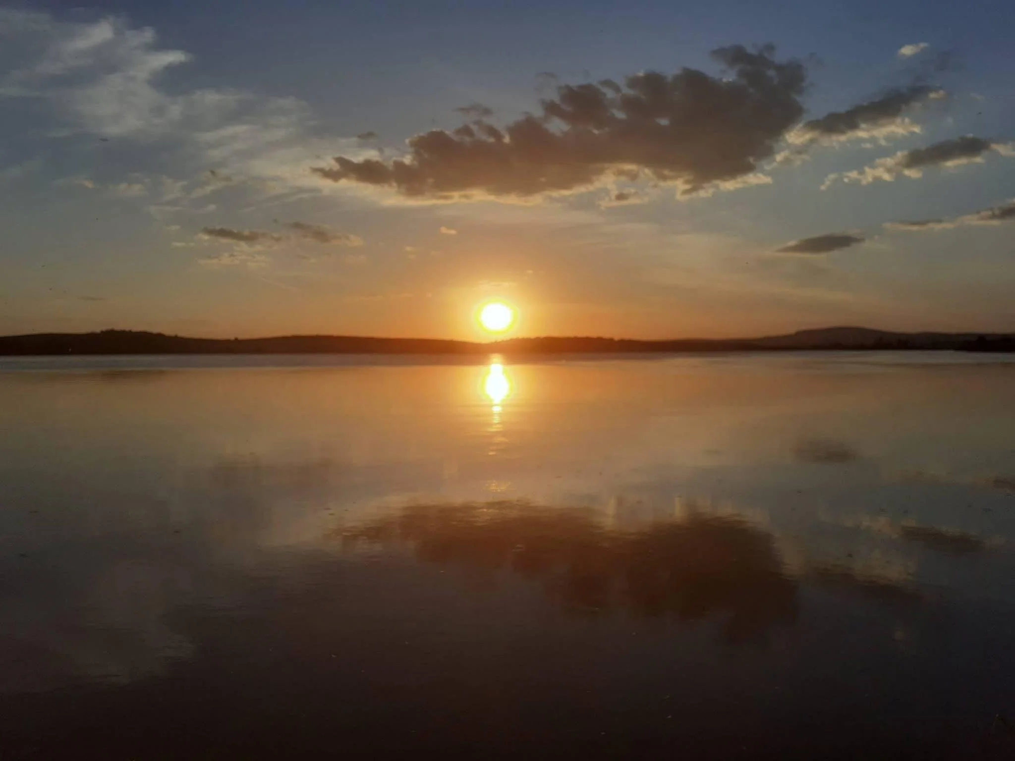 Sun setting over a calm body of water with scattered clouds in the sky.