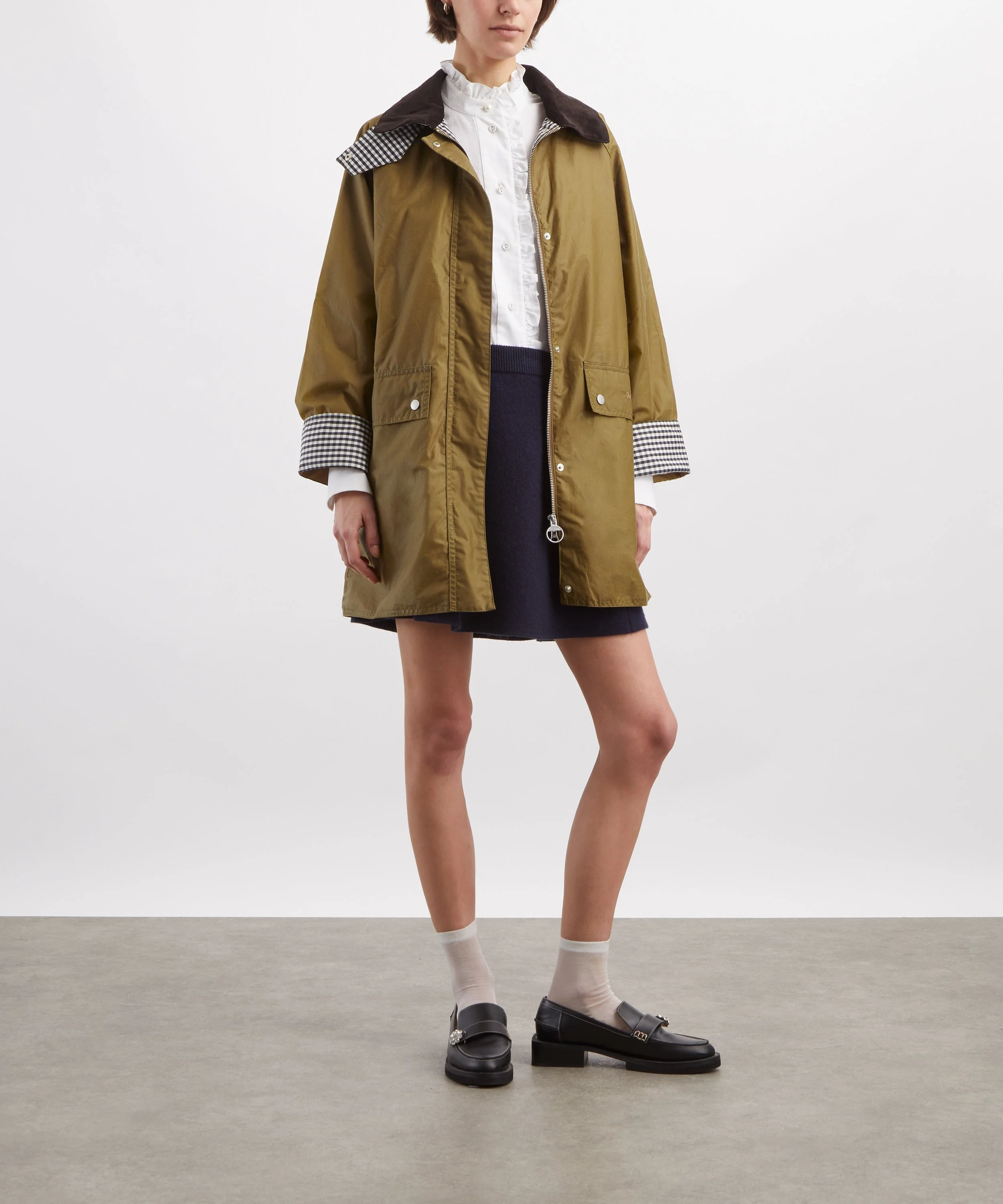 A woman standing with one leg slightly forward, wearing a brown raincoat with checkered cuffs, white shirt with ruffled collar, dark navy skirt, white socks, and black loafers against a plain white background.