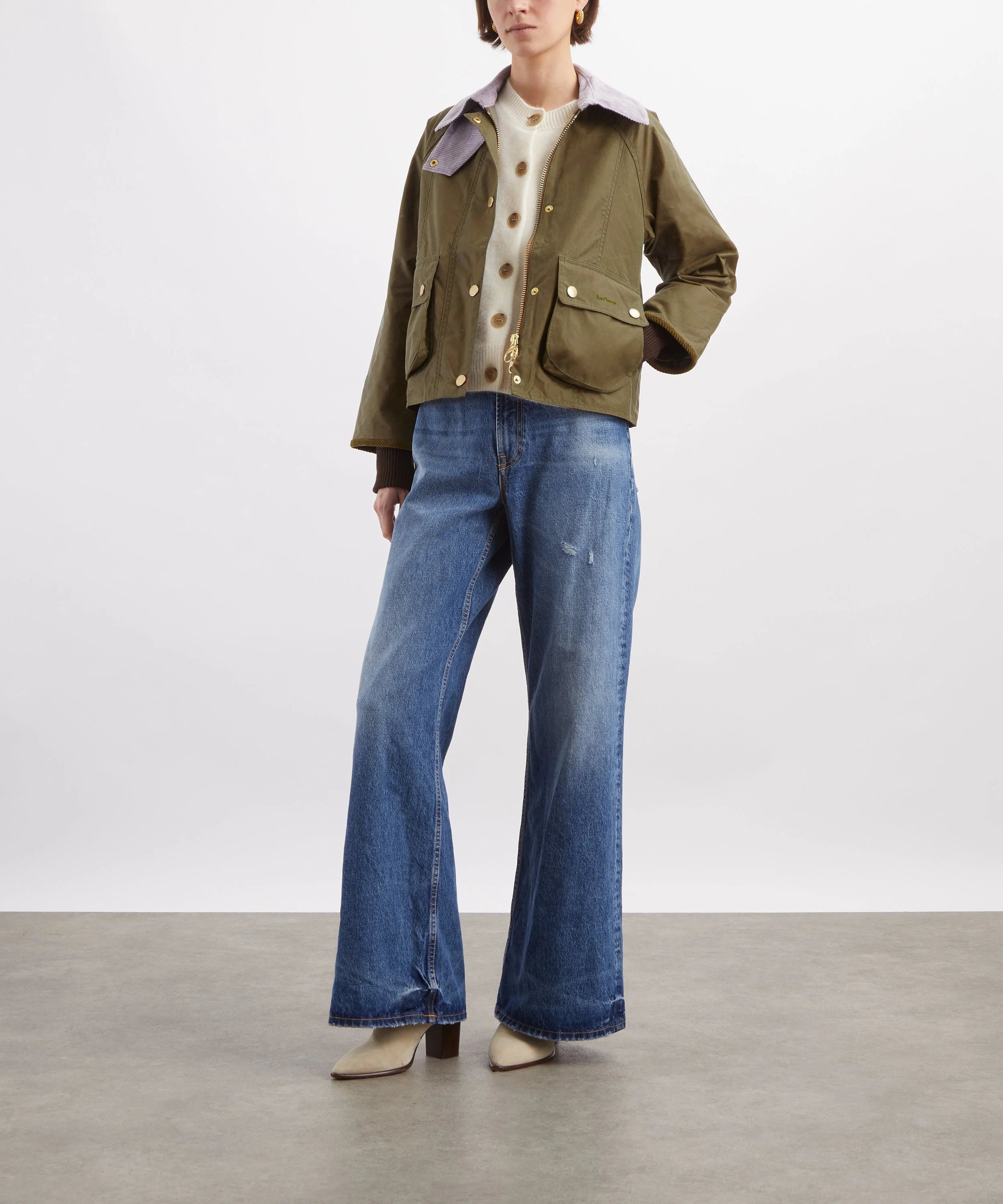 A woman wearing a brown jacket with purple collar, cream sweater with brown buttons, wide-leg blue jeans, and cream heeled boots, standing against a grey and white background.