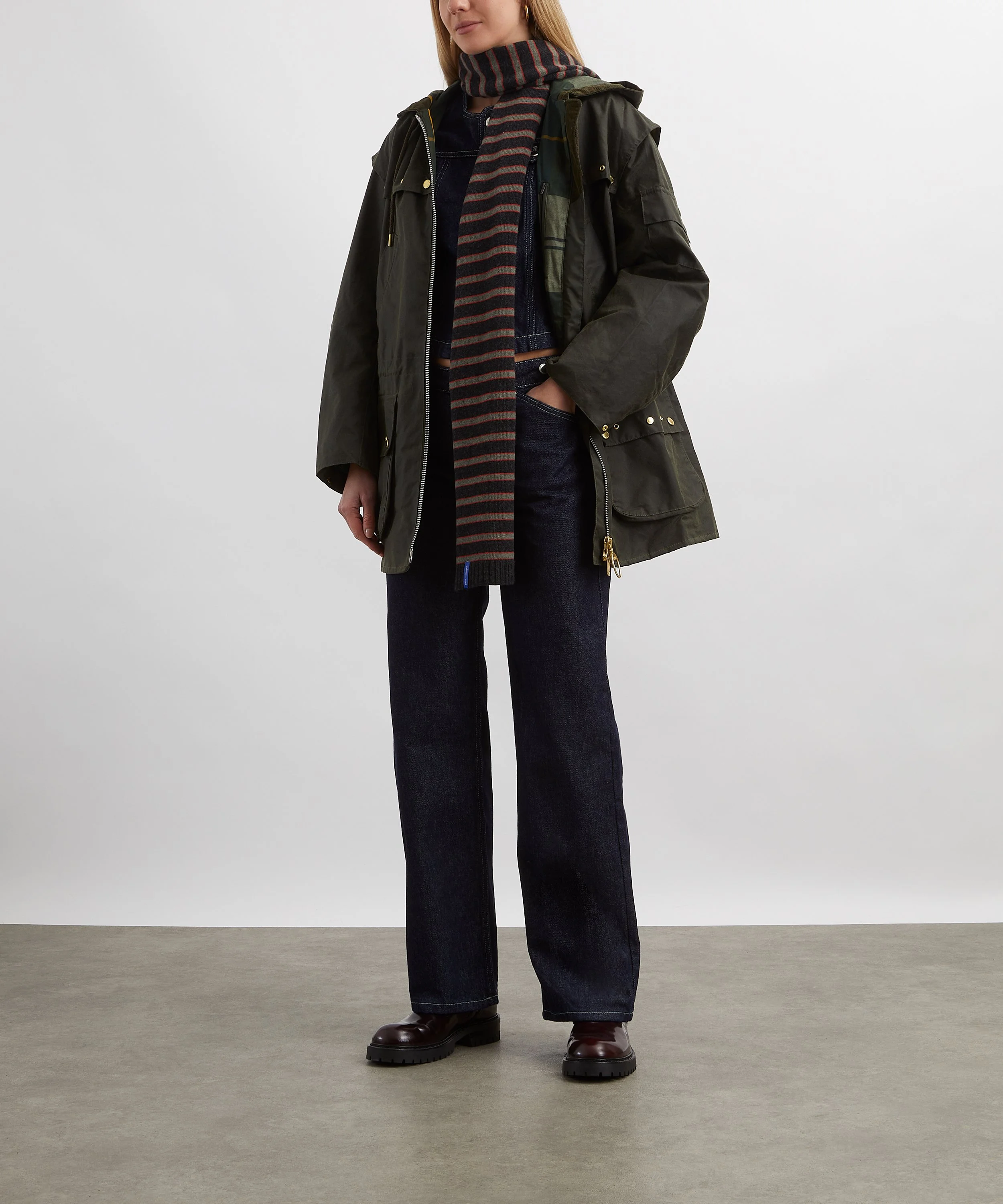 Person wearing a dark green jacket, dark jeans, brown boots, and a striped scarf, standing against a plain background.