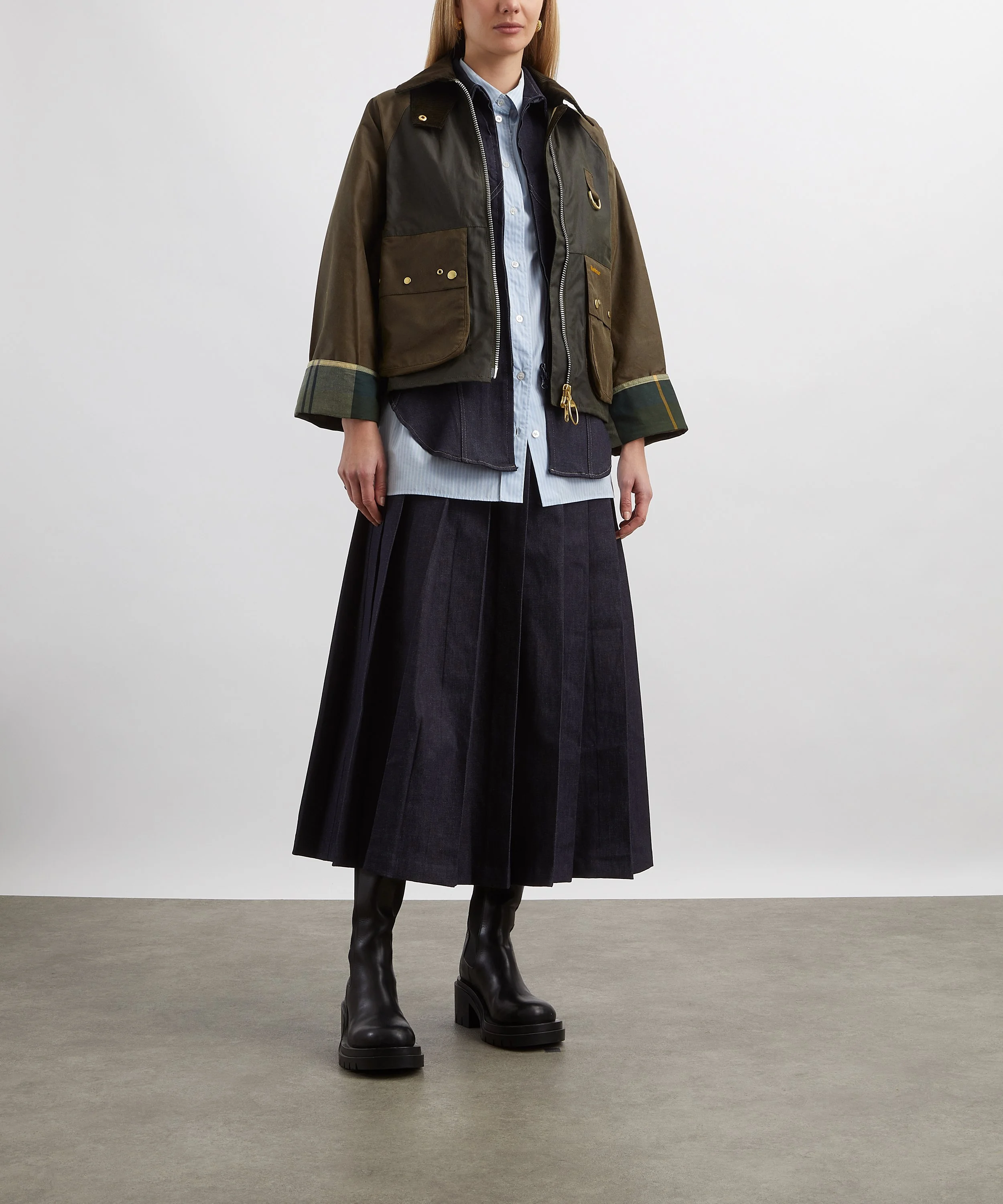 A woman wearing a layered outfit consisting of a dark-colored jacket over a denim shirt and black leather boots, standing against a plain background.