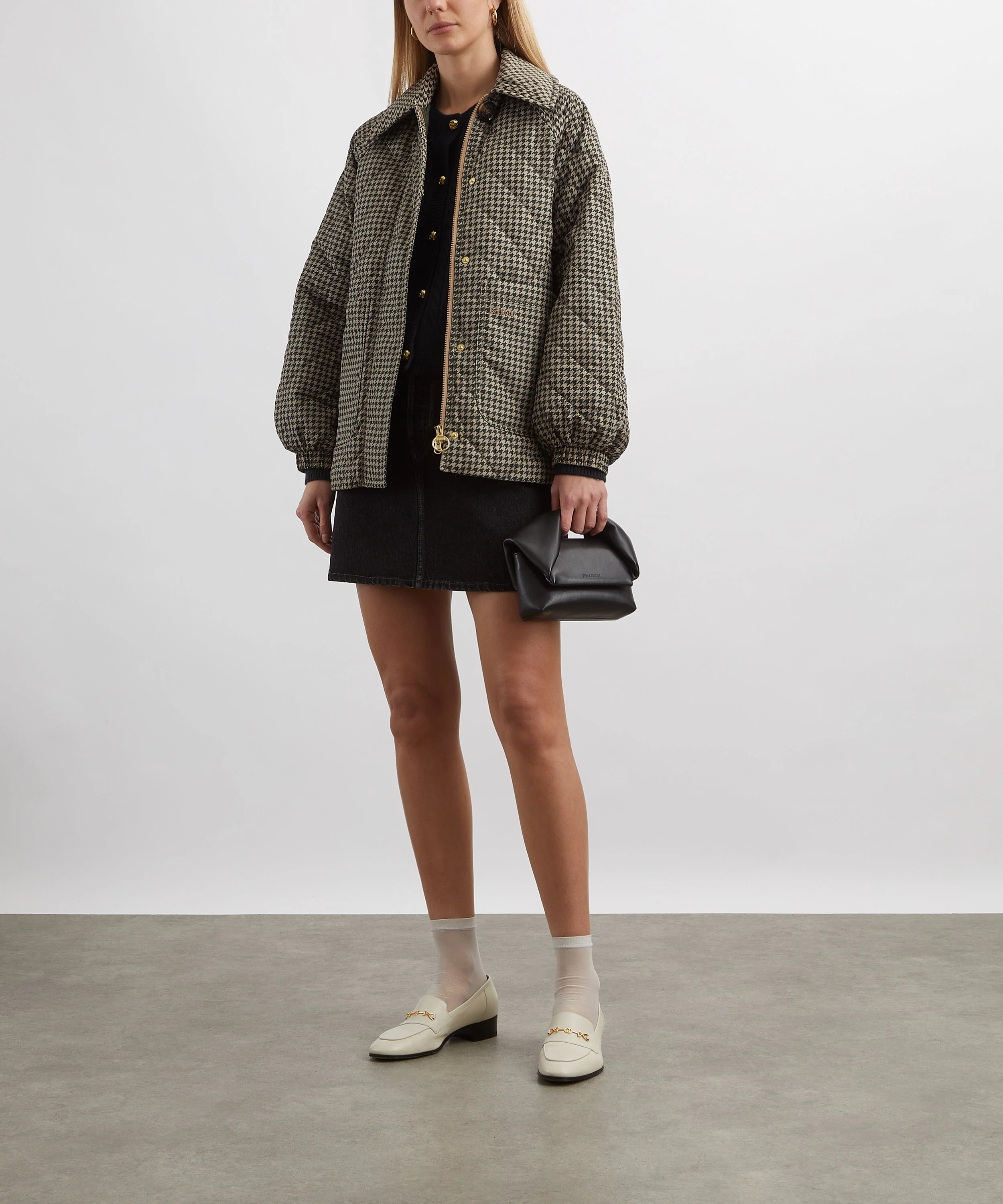 Person wearing beige and black houndstooth patterned jacket, black top, black skirt, white loafers with gold details, cream socks, holding a black clutch, standing against a plain background.