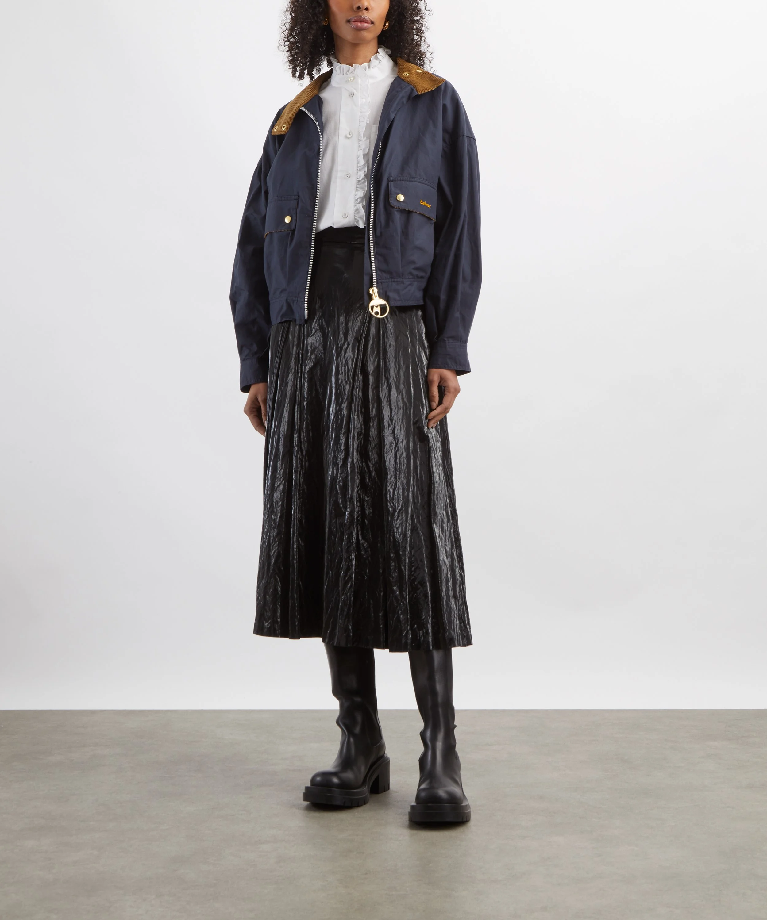 A woman standing with her lower half visible, wearing black platform boots, a black glossy skirt, and a navy blue jacket with brown accents over a white ruffled blouse against a plain white background.