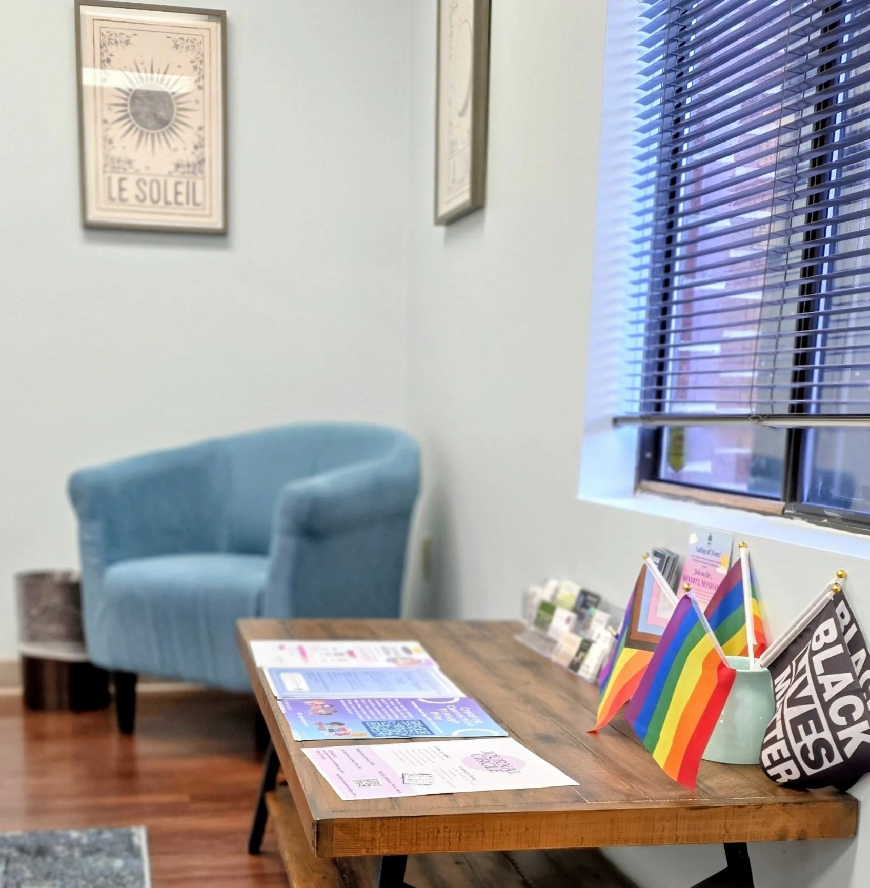 A waiting room with a light blue armchair, a wooden table with flyers and rainbow flags, a window with blinds, and framed sun-themed artwork on the light green wall.
