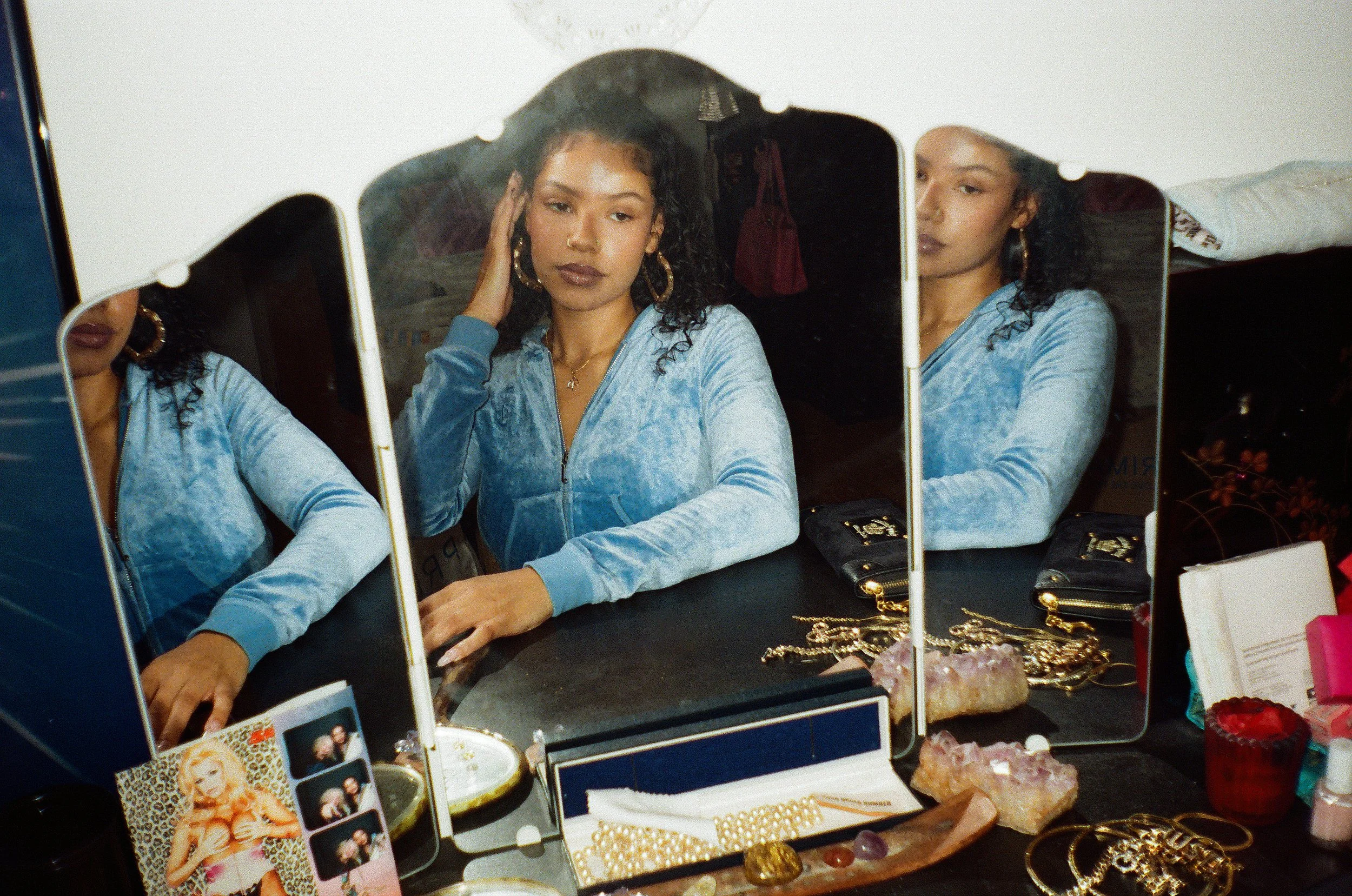 A woman sitting at a vanity table, looking at her reflection in a multi-panel mirror, wearing a light blue velour zip-up jacket, surrounded by jewelry, accessories, and beauty products.