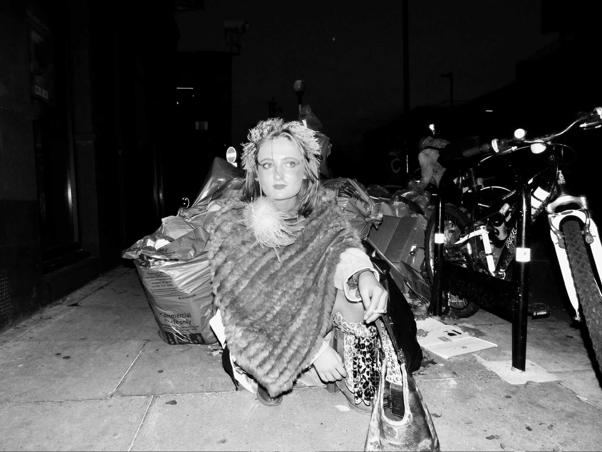 A person with short hair and makeup sitting on a shopping bag on a sidewalk at night, surrounded by bags and bicycles, with a dark street background.