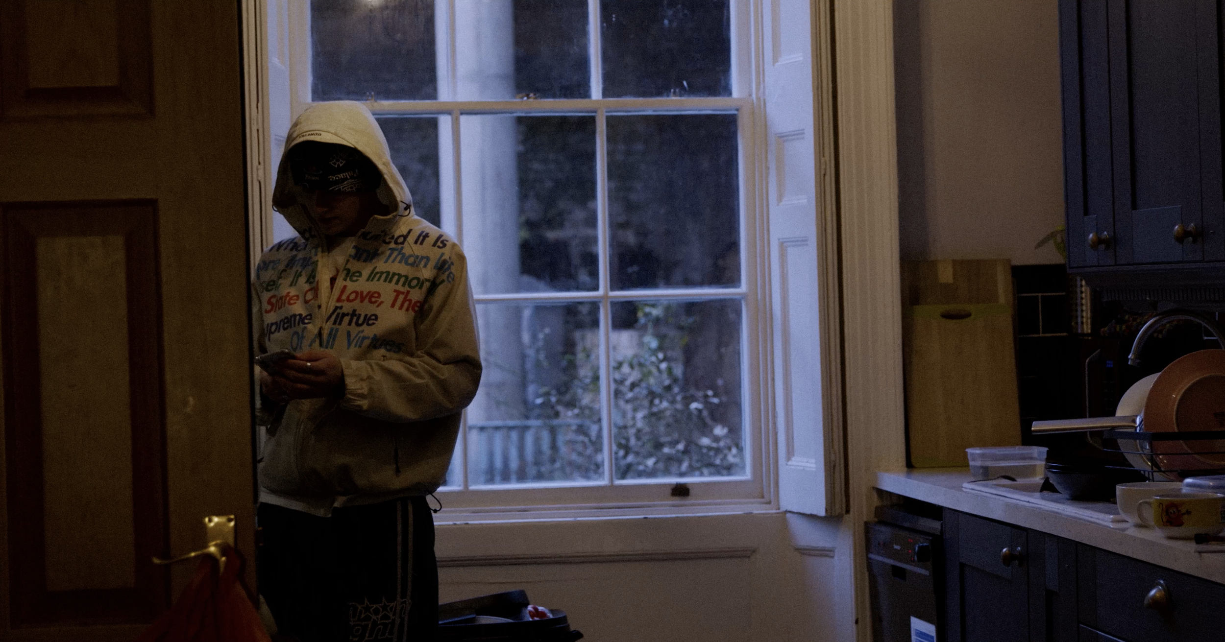 Person wearing a hoodie and sunglasses, looking at a phone in a dimly lit kitchen near a window.