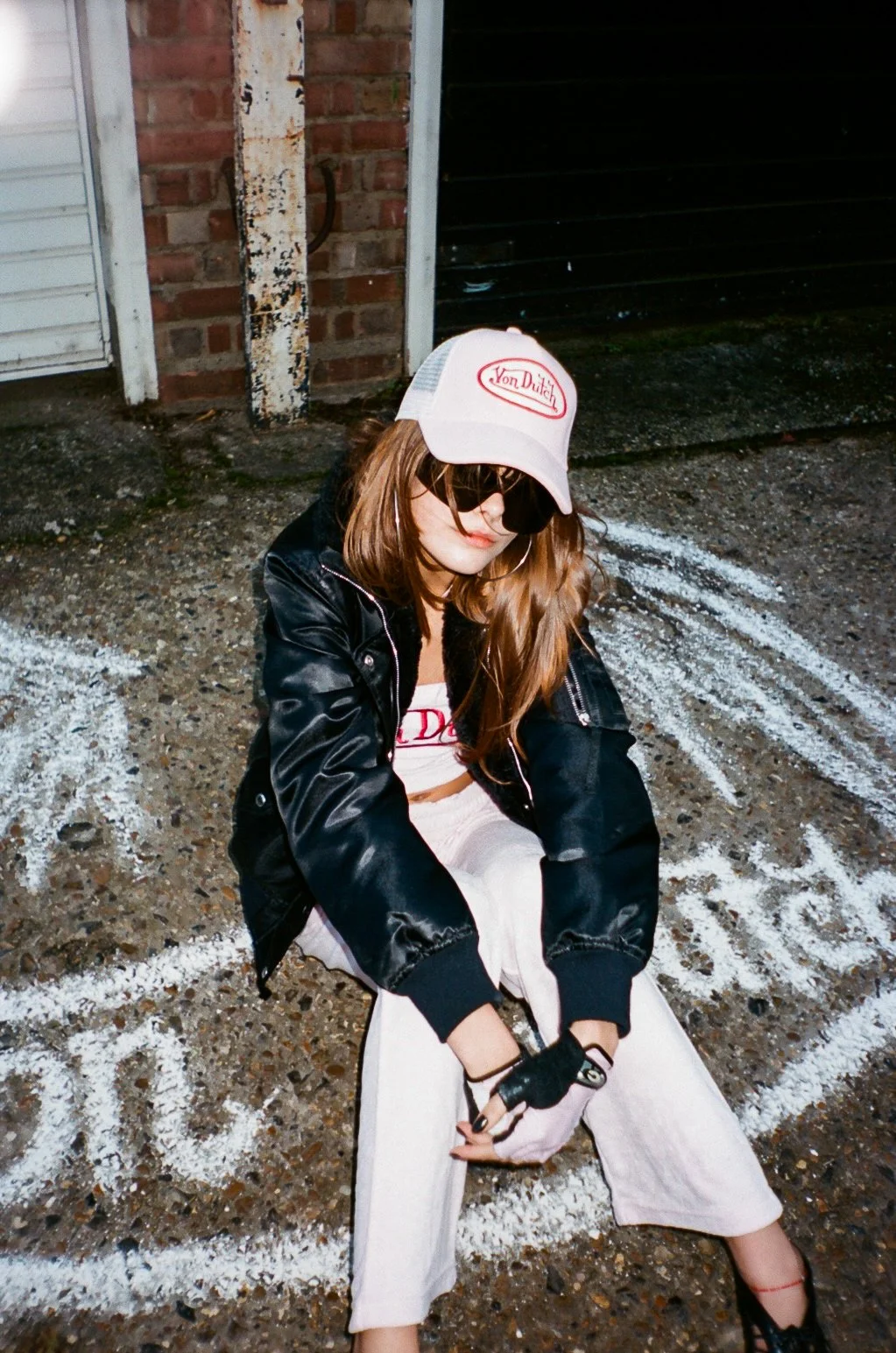 A young woman with long brown hair, wearing a white cap with red lettering, black sunglasses, a black leather jacket, a white crop top, and white pants, is sitting on the ground in front of a brick wall and black garage door with white chalk markings