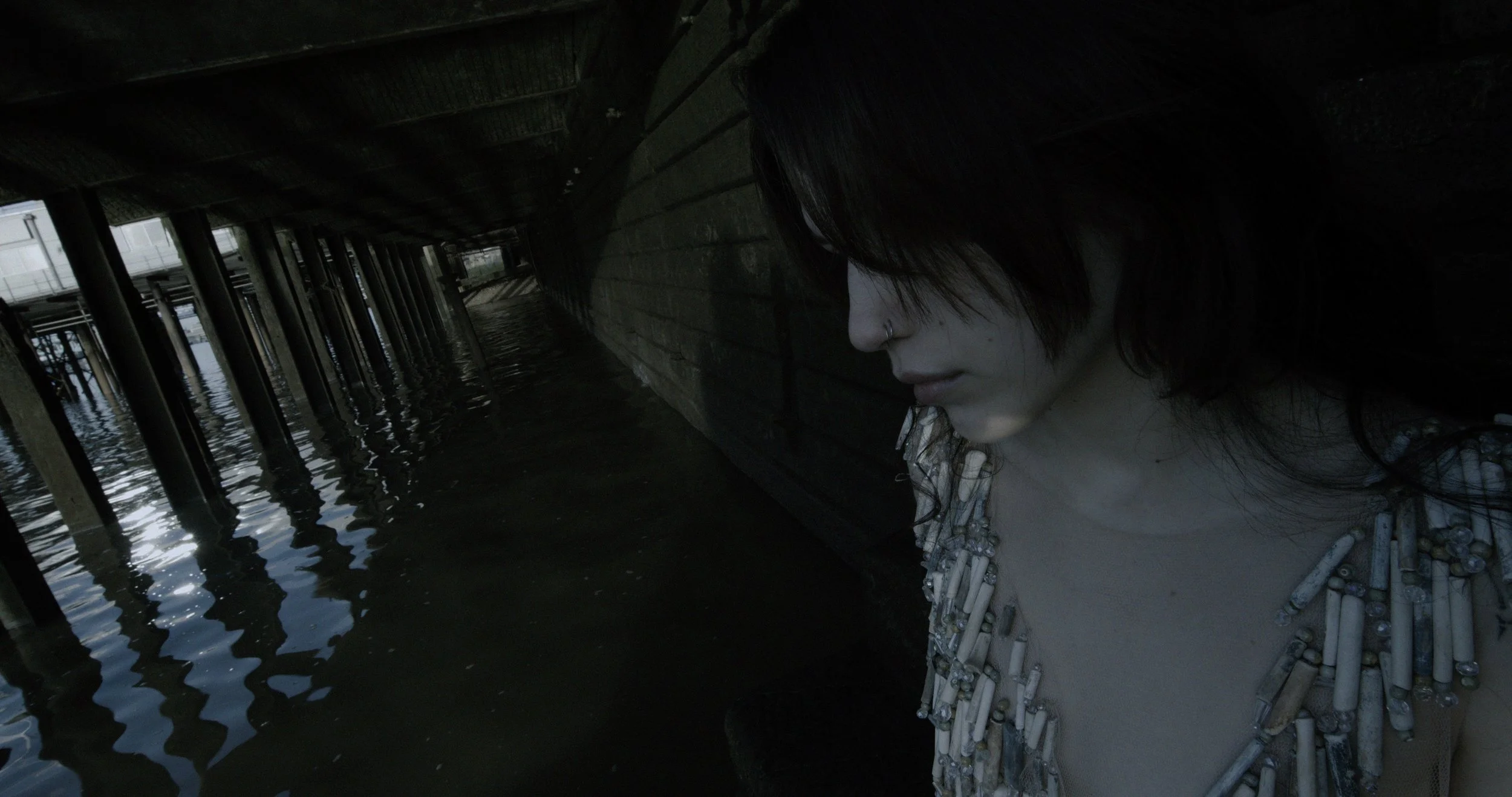 Person with dark hair and a nose piercing, wearing a top made of shells or beads, leaning against a wall under a pier near water with reflections of the pier's supports.