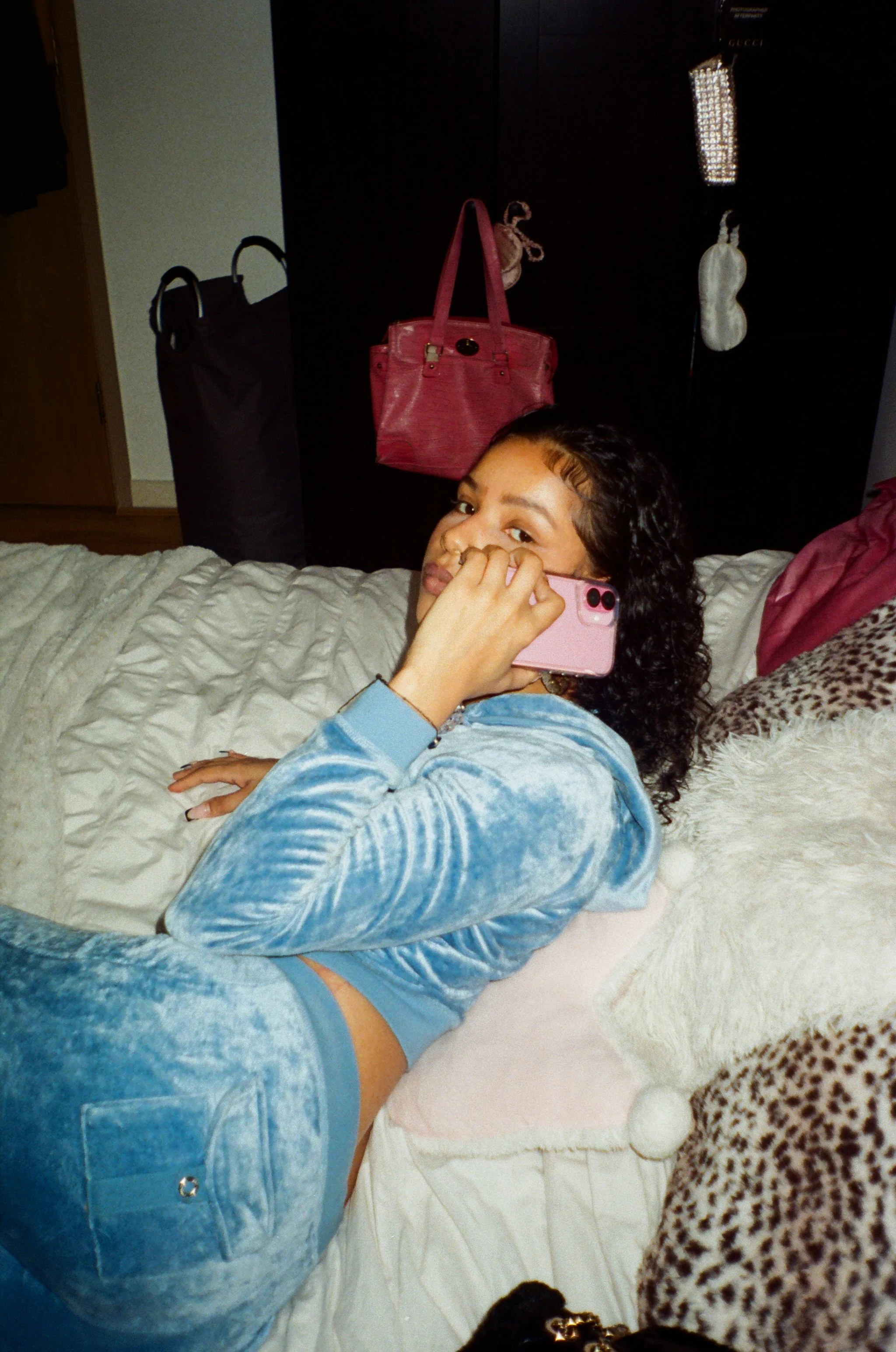 A young woman wearing a blue velvet tracksuit, is lying on a bed with white and leopard-print pillows, holding a pink smartphone to her ear, looking at the camera.