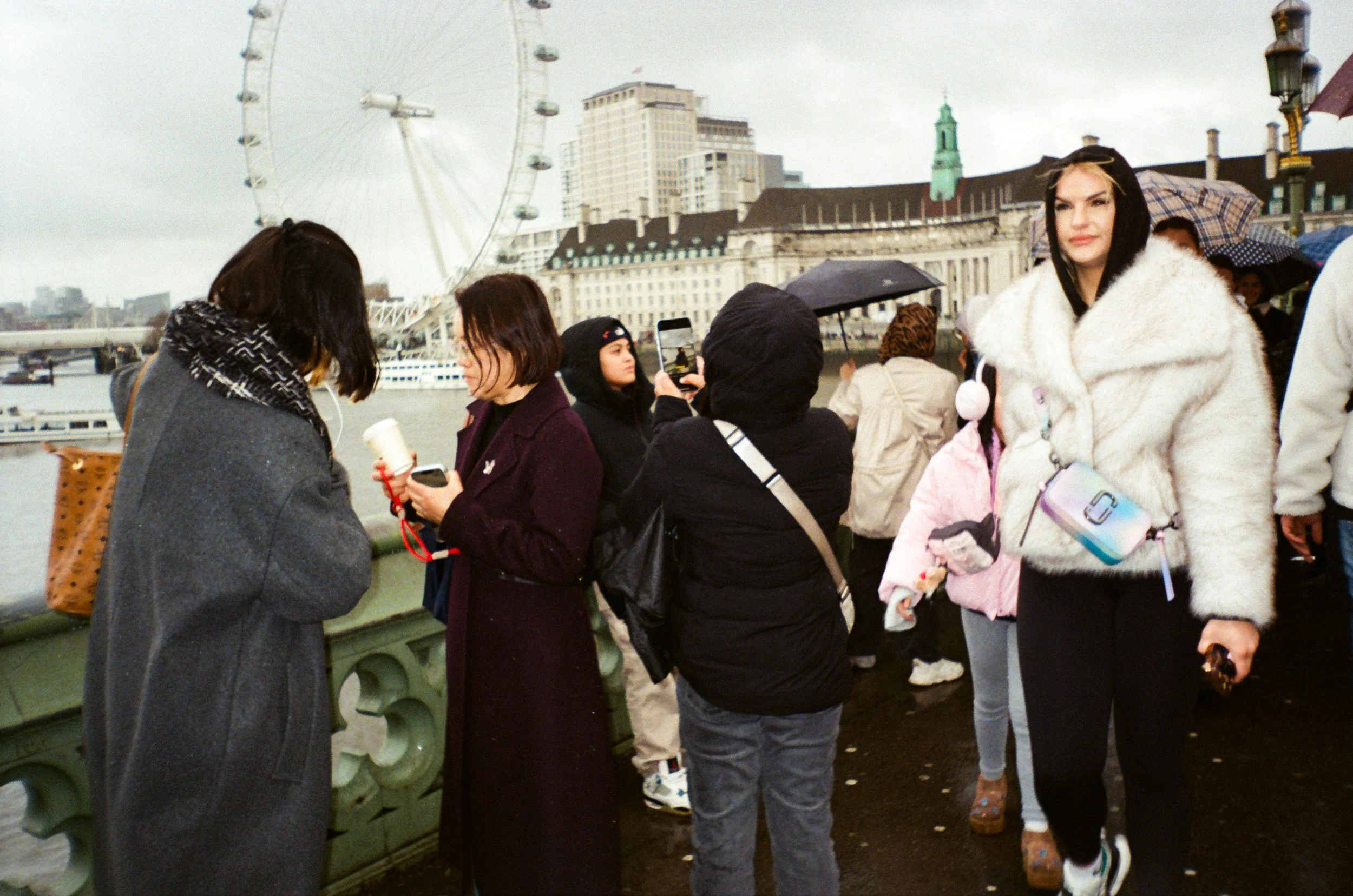 People standing on a bridge, some with umbrellas, with the London Eye Ferris wheel and city buildings in the background on a cloudy day.